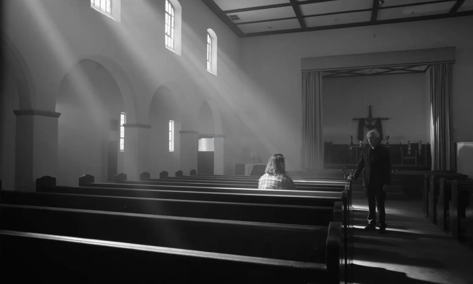 A church interior with sunlight streaming through windows, with two women, one sitting in church pews and the other standing, engaged in conversation near the altar.