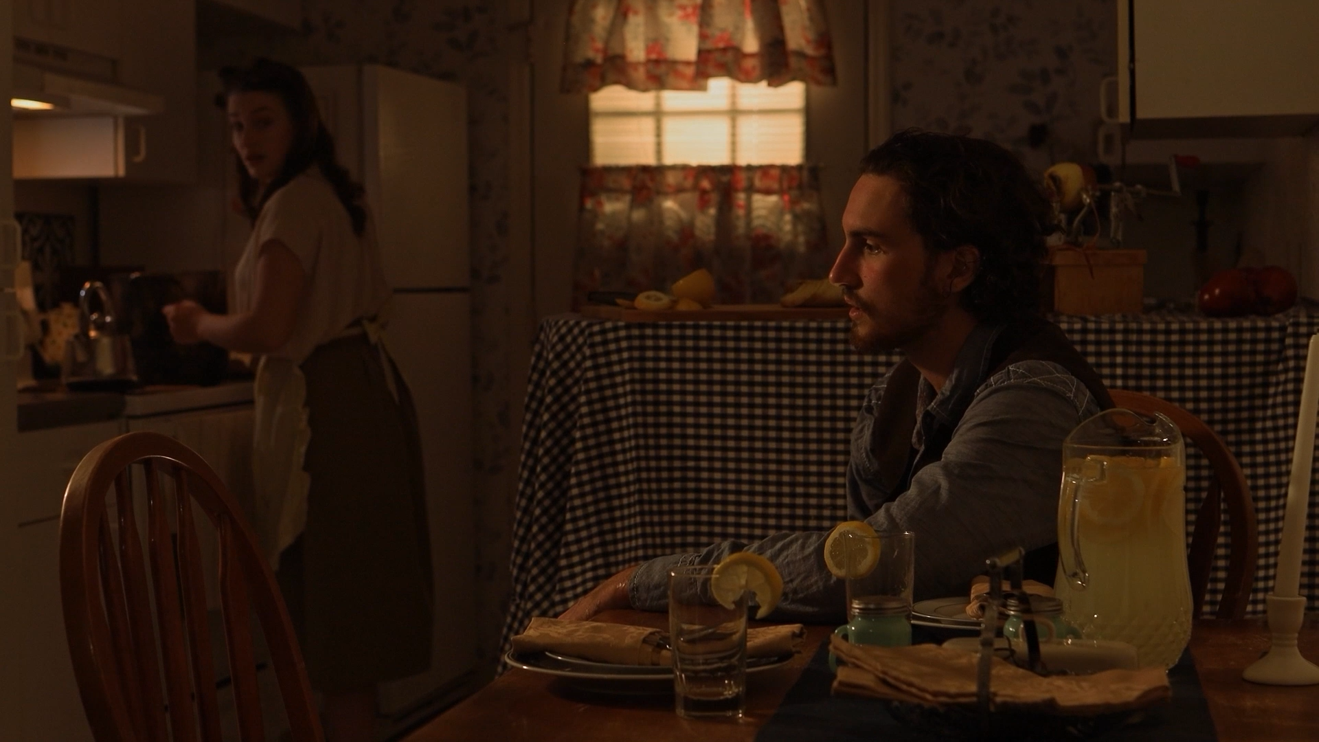 A man sitting at a dining table with lemon water, a pitcher of lemonade, and a plate with a napkin, with a woman standing in the kitchen in the background.