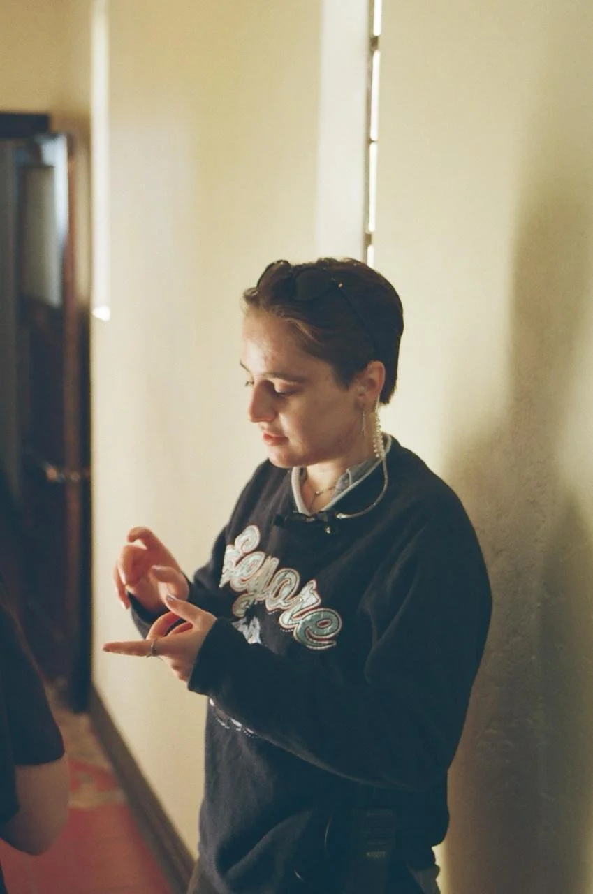 A young woman with short hair, wearing a black sweatshirt, is standing indoors near a wall. She is looking at her phone and appears to be focused or engaged. She's also wearing glasses pushed up on her head, earrings, and a necklace.