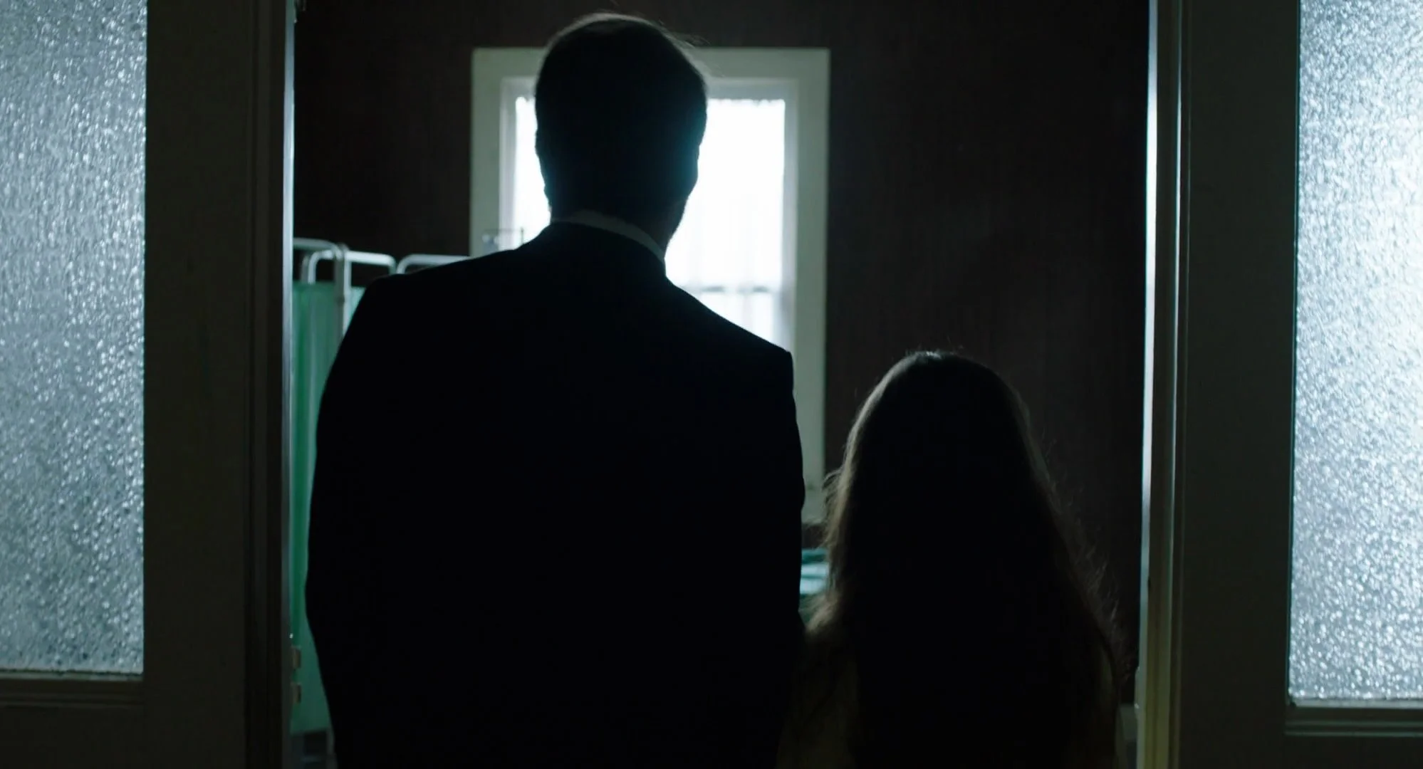A man and a young girl standing in a dimly lit room, facing a window with light shining through, with hospital beds partially visible.