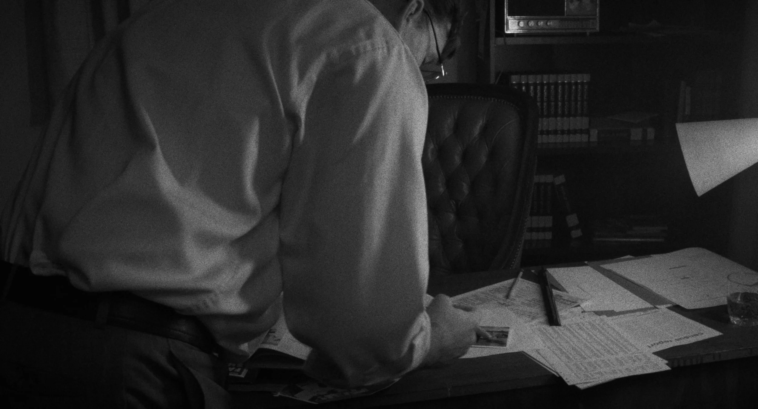 A man in a light-colored shirt is sitting at a desk, signing a photograph. The desk is cluttered with papers, a pen, and a glass of water. There are bookshelves and a stereo in the background.