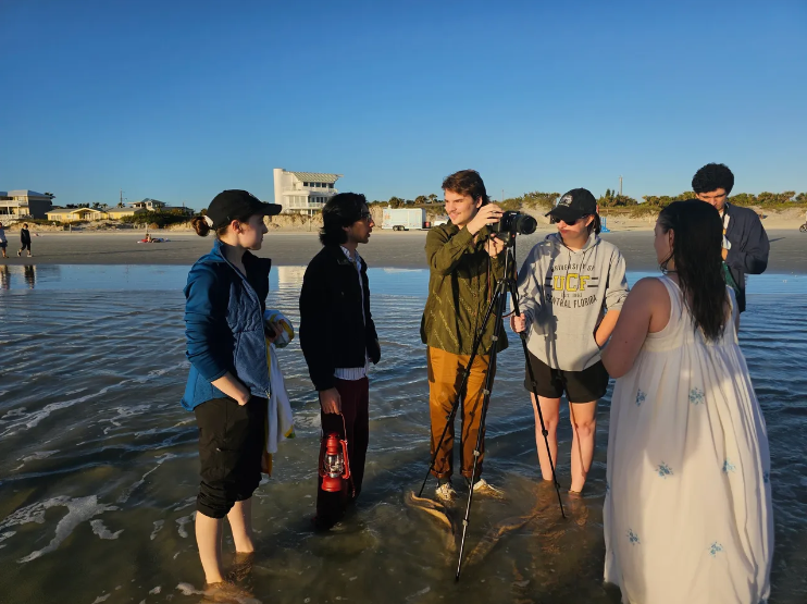 A group of six people on the beach during sunset, with five of them being interviewed or taking photos of a woman in a white dress, while a photographer takes pictures. The background shows the ocean, sandy beach, and buildings in the distance.