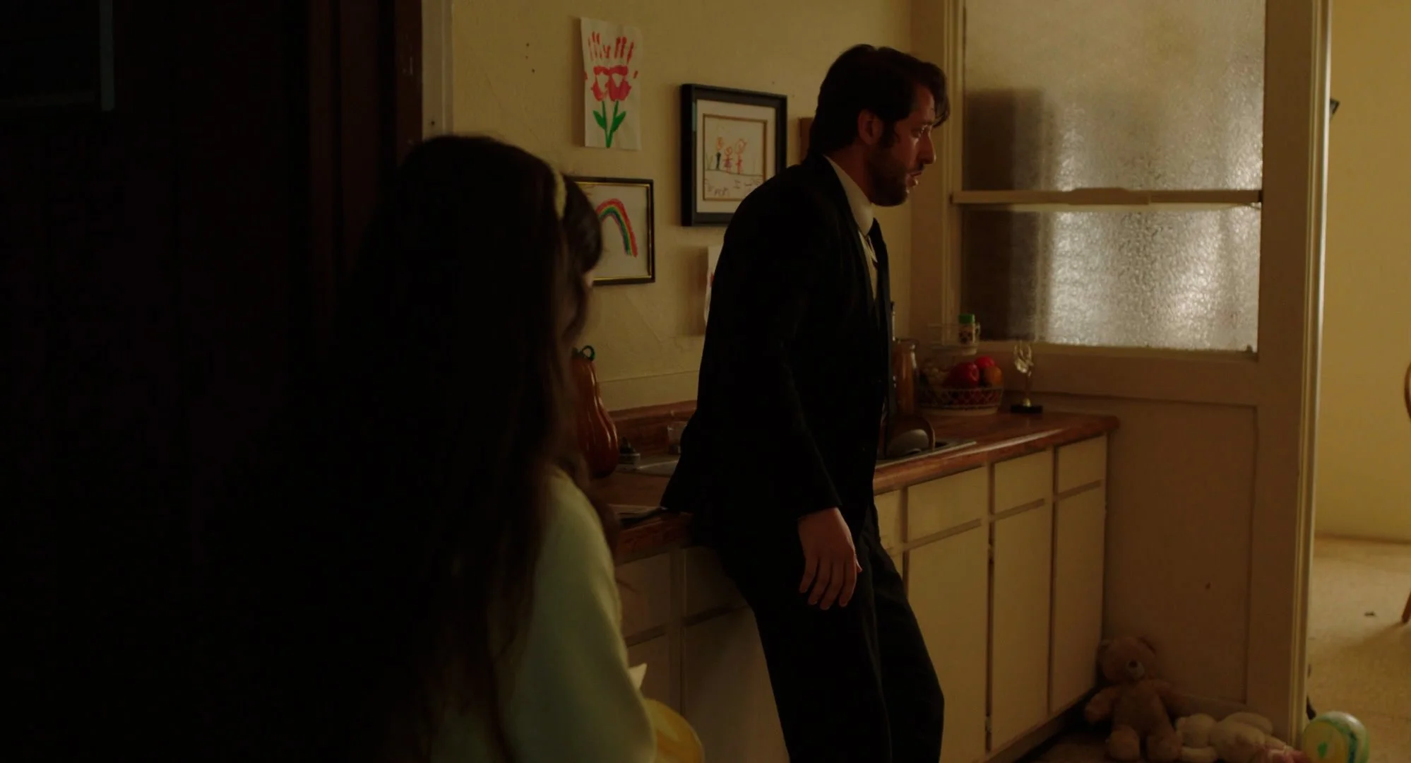 A man in a suit leaning against a kitchen counter, with a woman partially visible in the foreground. The kitchen wall displays children's artwork, including a flower, rainbow, and a drawing. The scene is dimly lit with a warm glow.