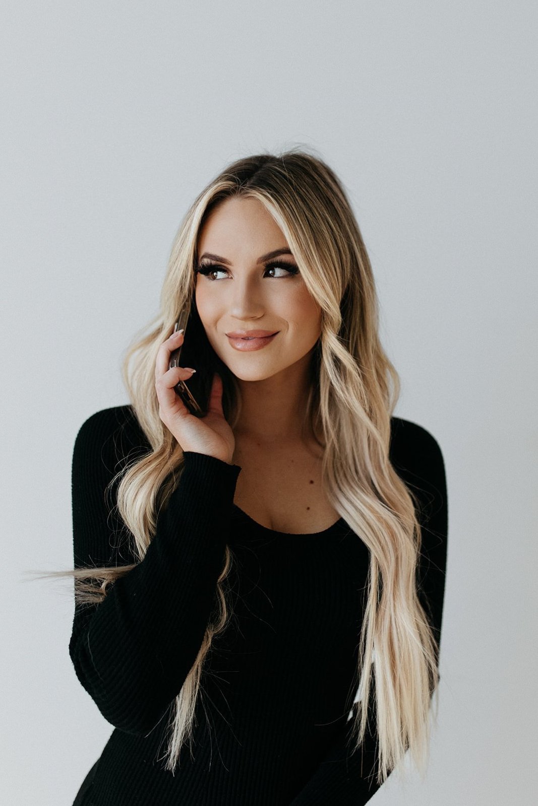 Young woman with long blonde hair and makeup talking on her phone, wearing a black top, against a plain white background.