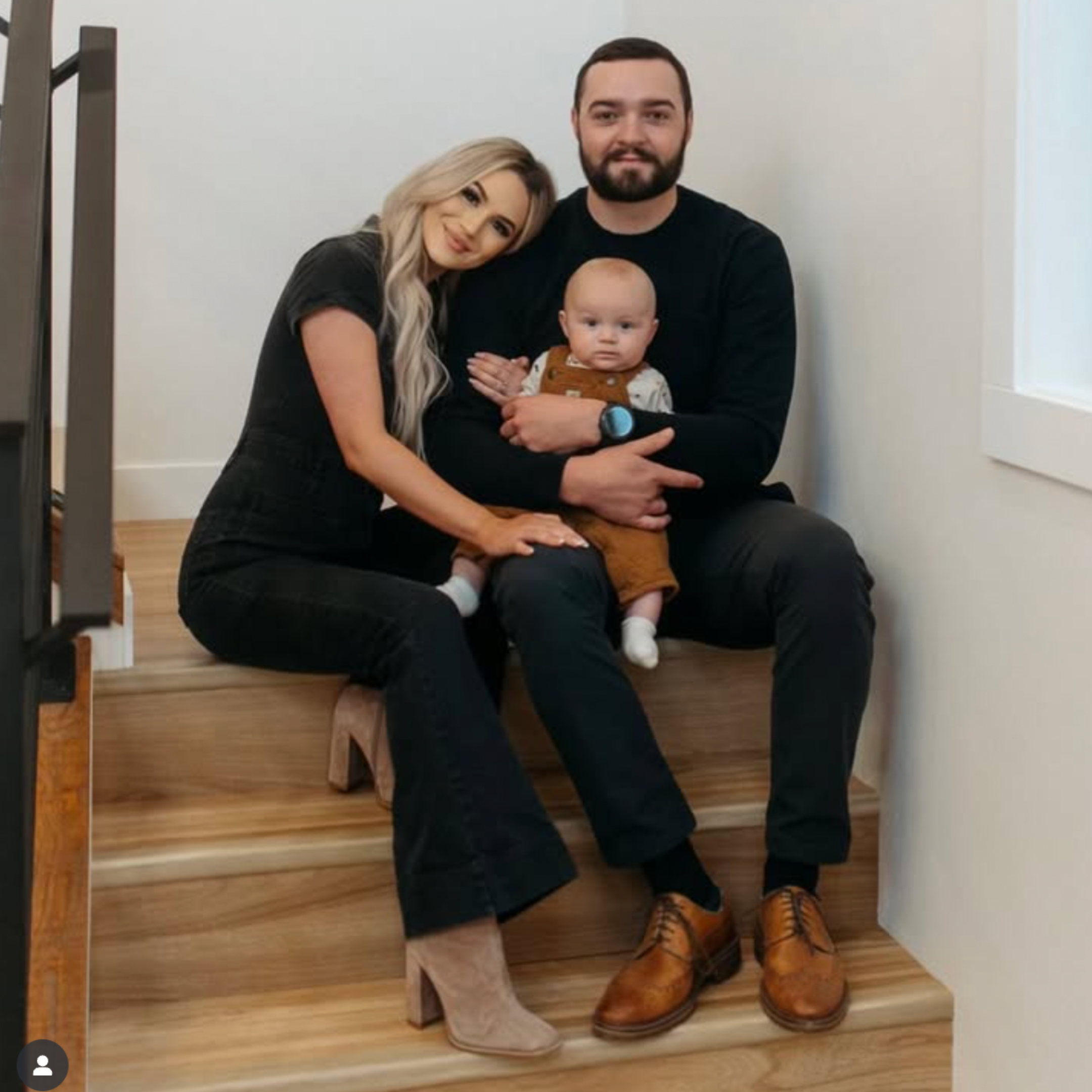 A family of three sitting on wooden stairs, with a woman and a man holding a baby. The woman has long blonde hair and is wearing a black outfit with beige boots. The man has a beard, short dark hair, and is wearing a black shirt with brown shoes. The baby has light hair and is dressed in a brown outfit with white socks.