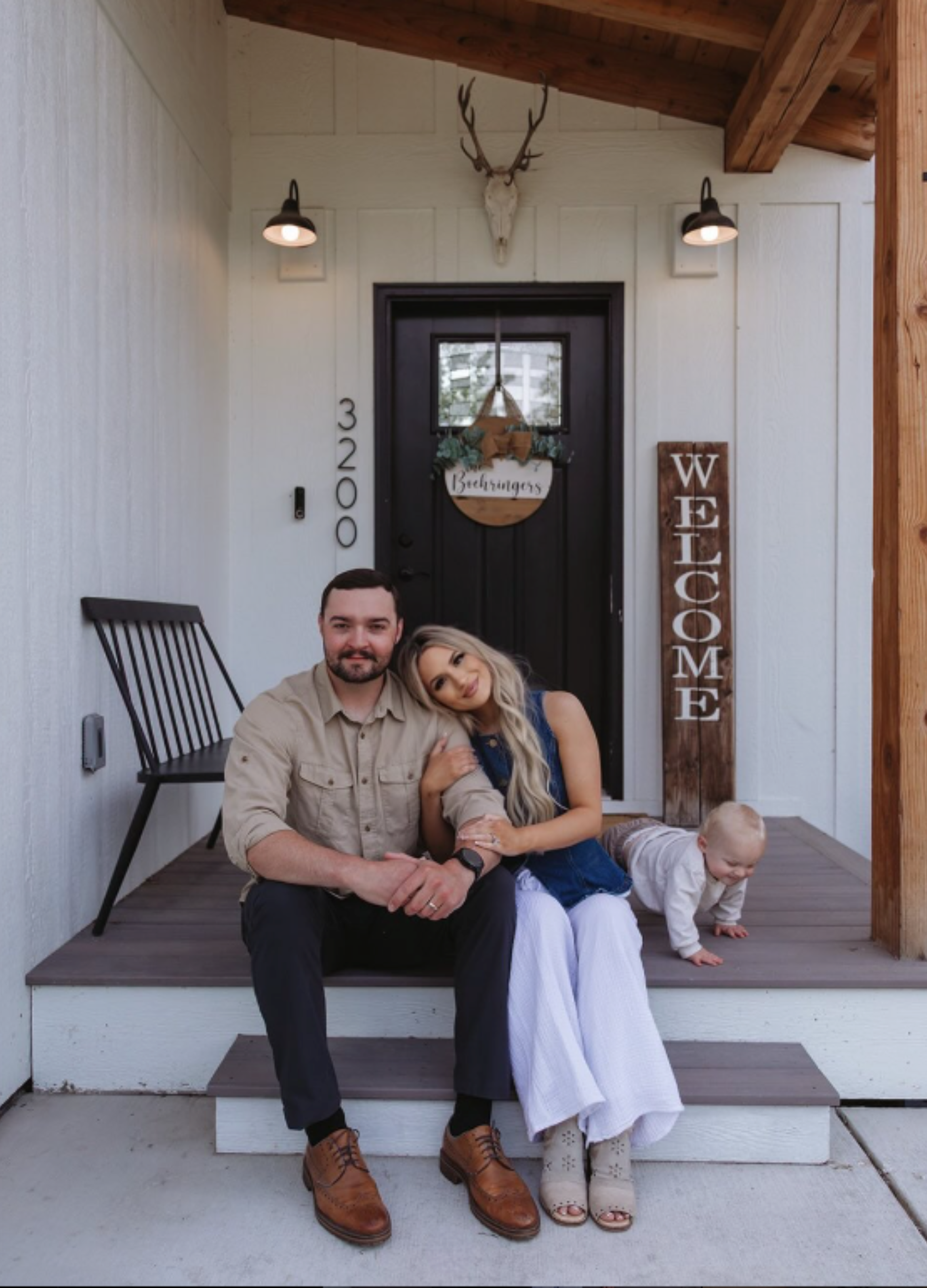 A family of three sitting on the front porch of a house. The man and woman are seated, with the woman leaning her head on the man's shoulder. The boy is crawling on the porch behind them. The house has a black door with a decorative sign, a vertical 'WELCOME' sign, and a deer head mounted above the door. The porch has wooden steps and a wooden roof.