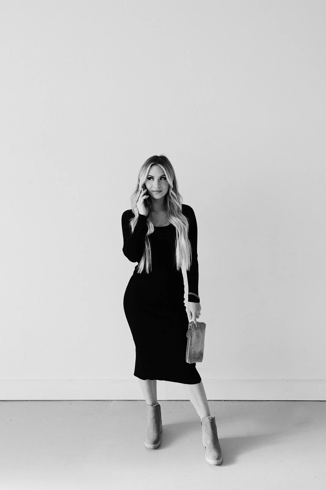 Black and white photo of a woman standing against a plain wall, holding a phone to her ear with one hand and a clutch purse in the other, wearing a long-sleeved dress and high-heeled boots.