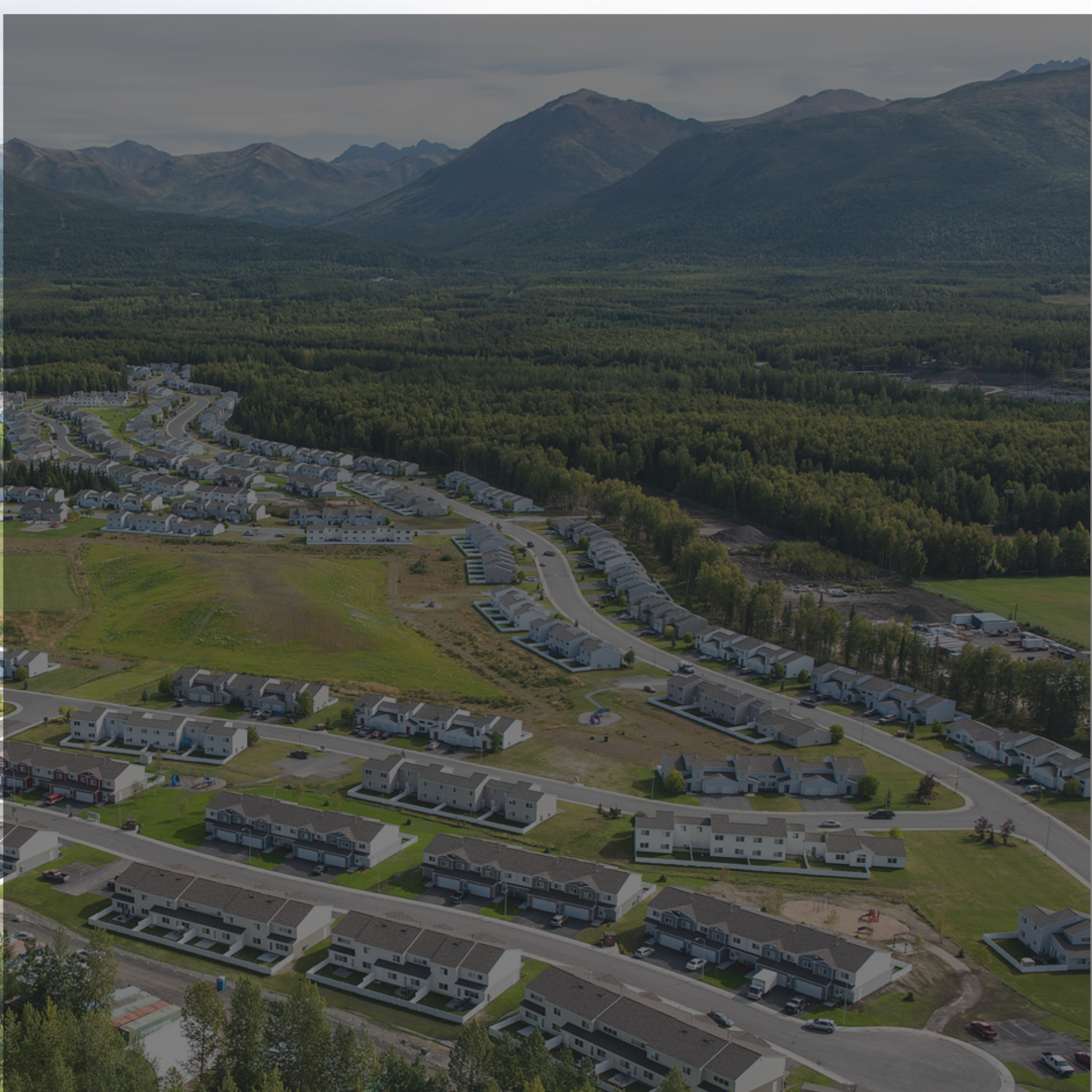 Aerial view of a suburban neighborhood with rows of houses, green lawns, and winding roads, surrounded by a forest and mountains in the background.