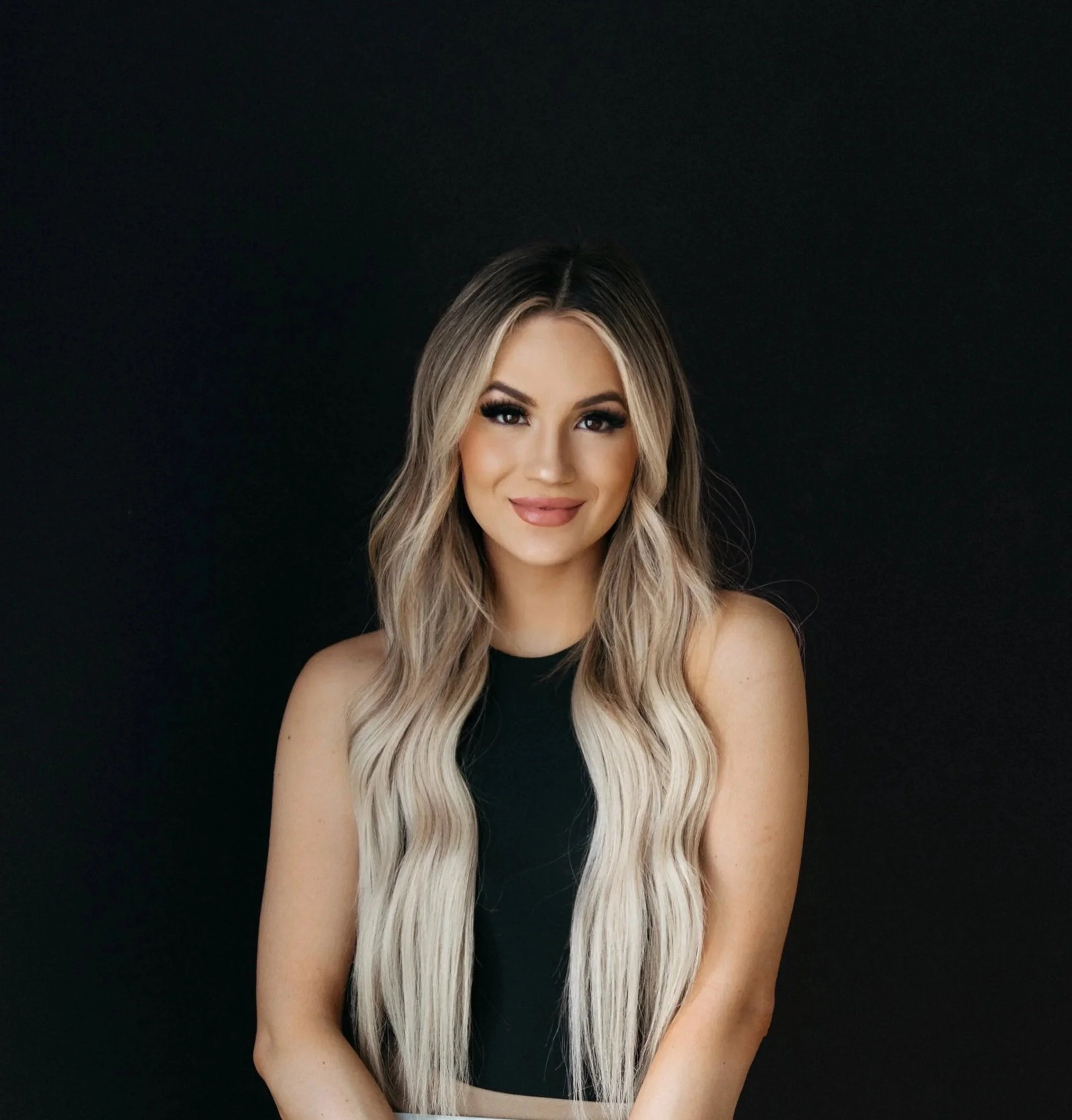 A young woman with long, wavy blonde hair, wearing a black sleeveless top, smiling against a dark background.