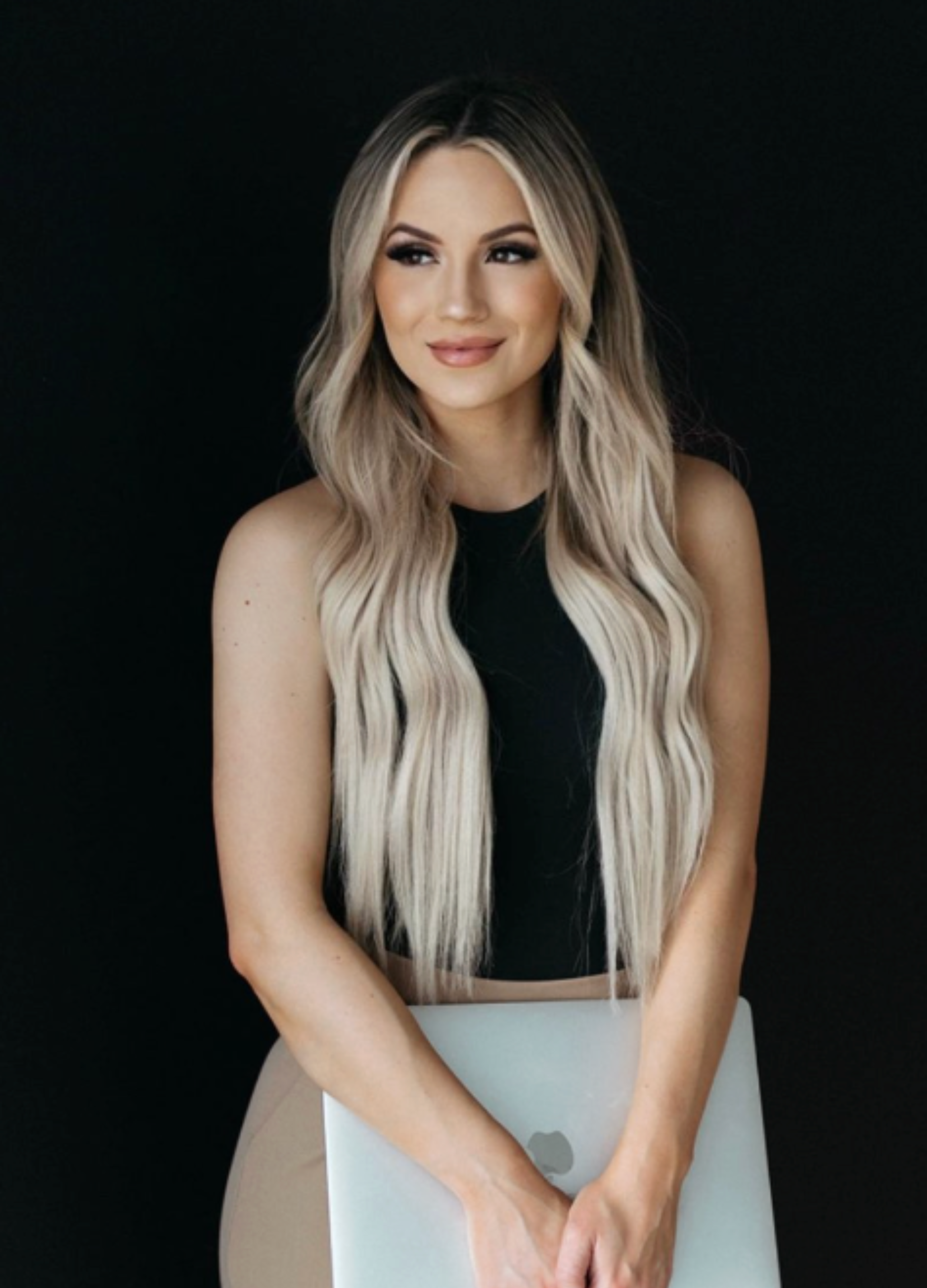 A woman with long, wavy blonde hair, wearing a black sleeveless top, holding a silver MacBook, and smiling softly against a black background.