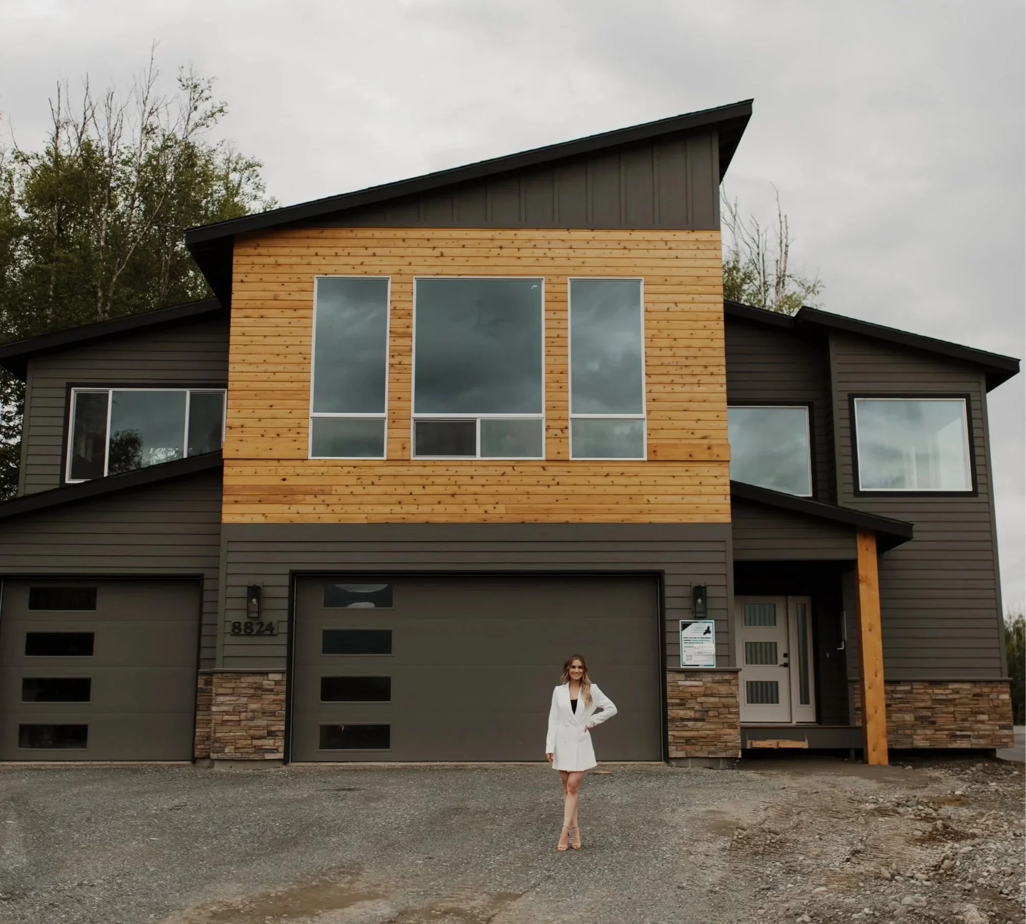 A modern multi-story house with gray and wooden exterior, large front windows, and a woman in a white dress standing near the garage entrance.