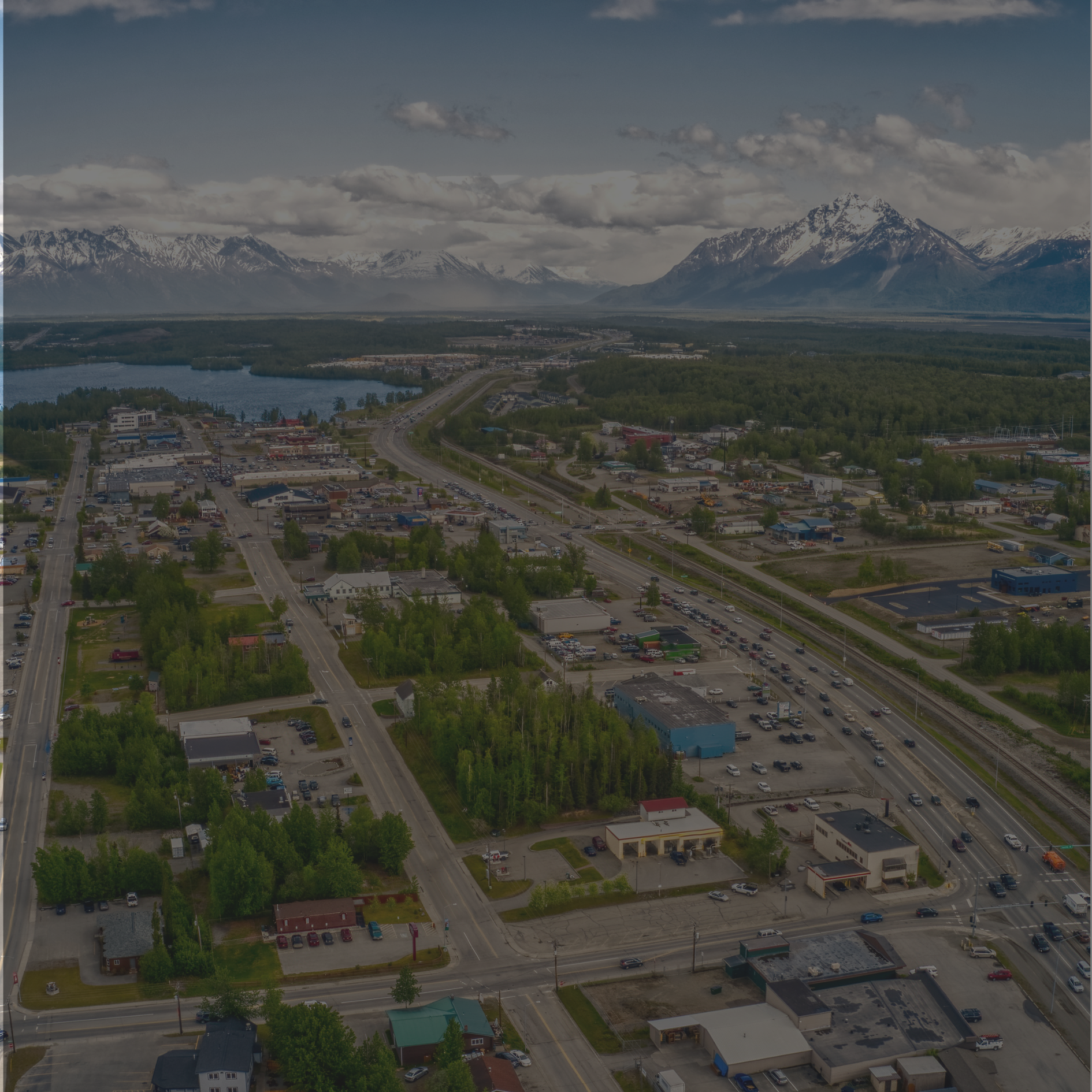 Aerial view of a small town with commercial buildings, roads, and parking lots, surrounded by green trees and backed by snow-capped mountains and a lake in the distance.