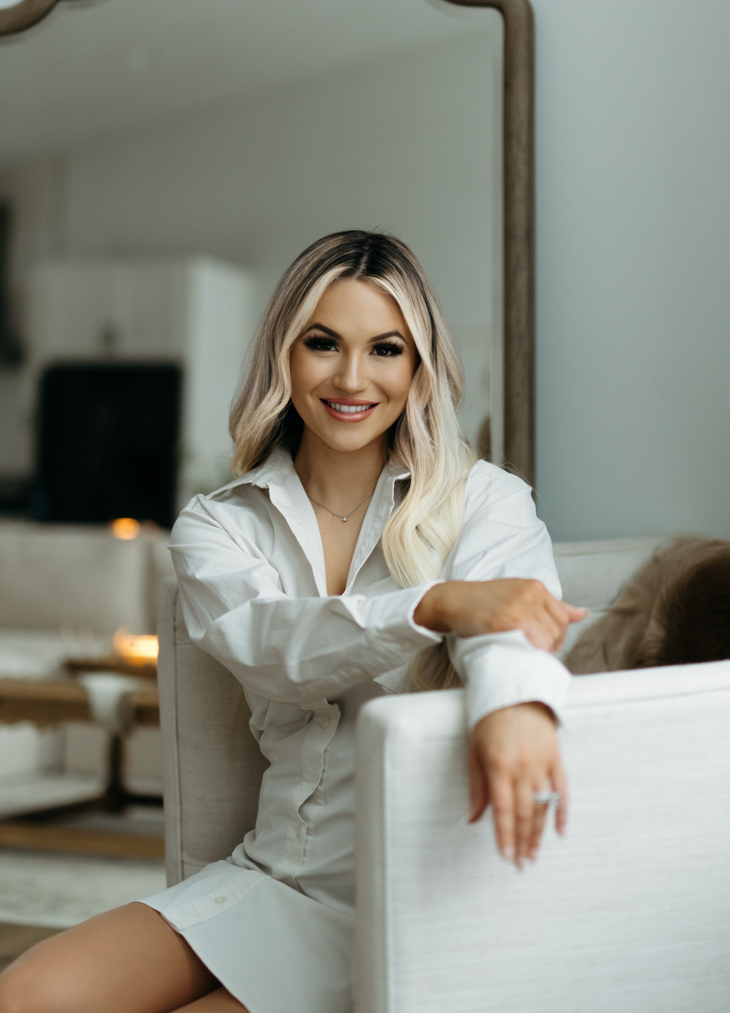 A woman with long blonde hair, wearing a white shirt, sitting on a beige sofa, smiling at the camera in a well-lit living room.