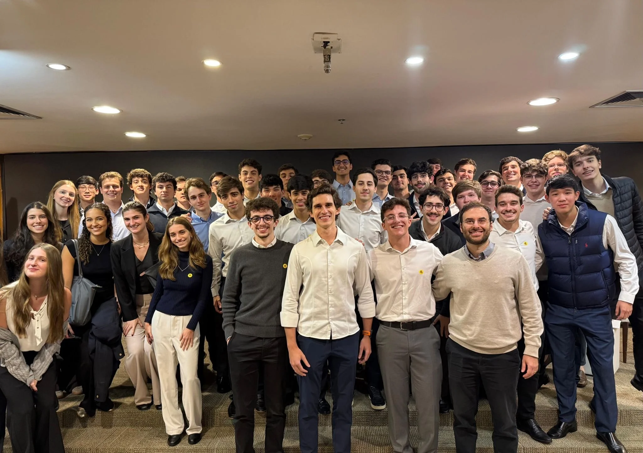 Grupo de jovens sorrindo em foto de frente, em ambiente interno com iluminação de teto.