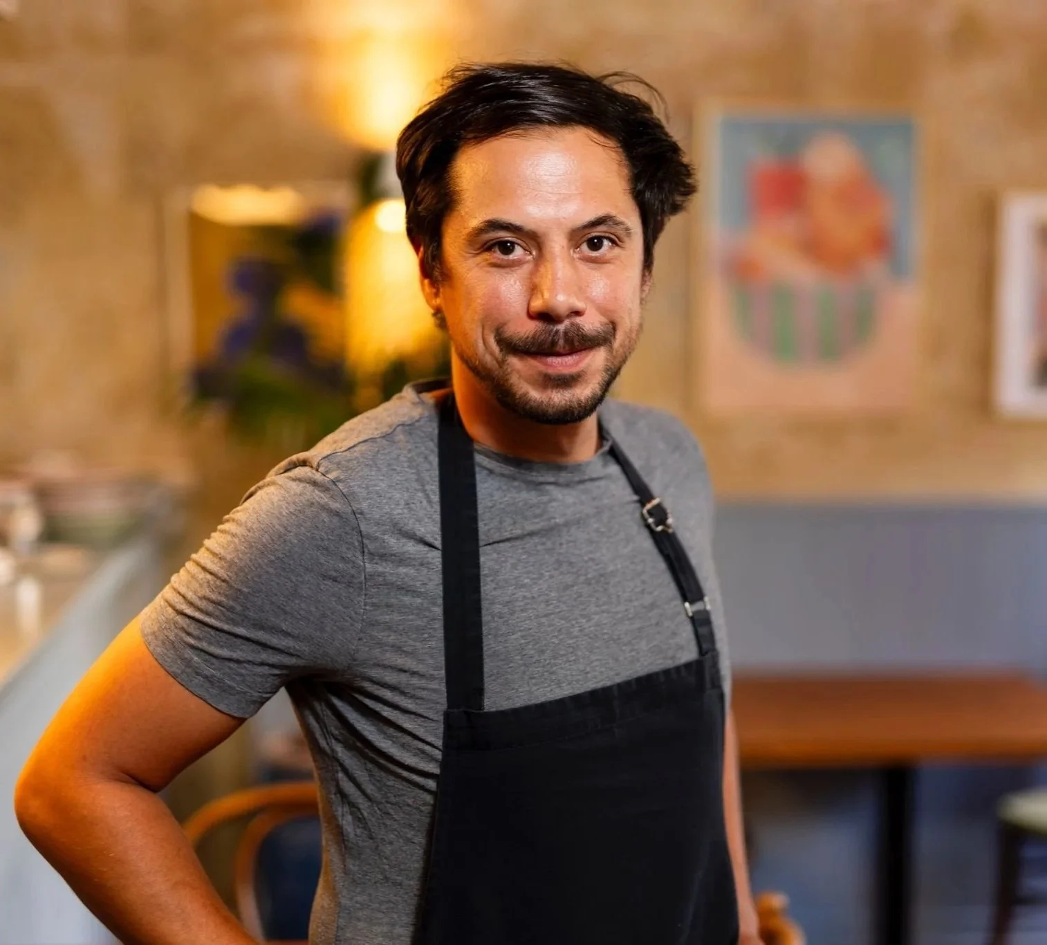 A man with dark hair and a beard wearing a gray T-shirt and a black apron, smiling in a warmly lit indoor setting, possibly a restaurant or a kitchen.