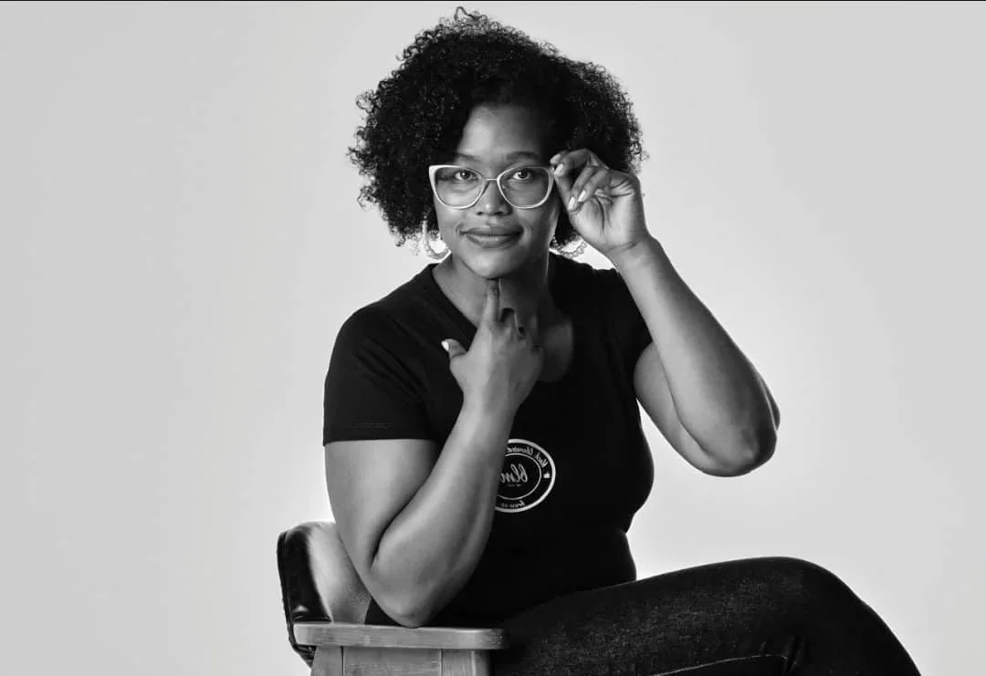 Black woman with curly hair wearing glasses sitting on a chair, holding her glasses and touching her neck, looking at the camera.