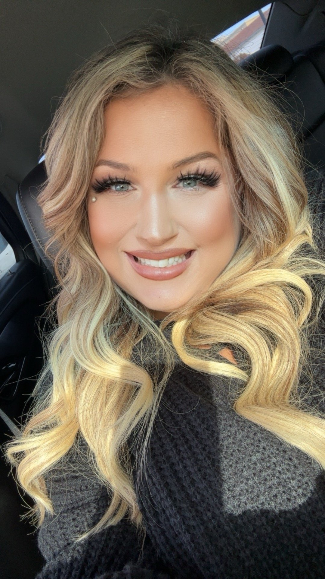 Close-up selfie of a smiling woman with blonde, wavy hair and bright blue eyes, sitting inside a car.