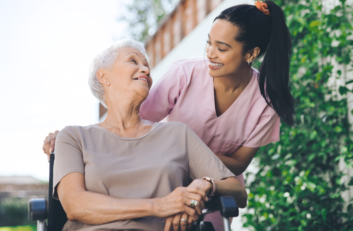 A young woman wearing pink scrubs smiling and looking at an elderly woman in a wheelchair outdoors.