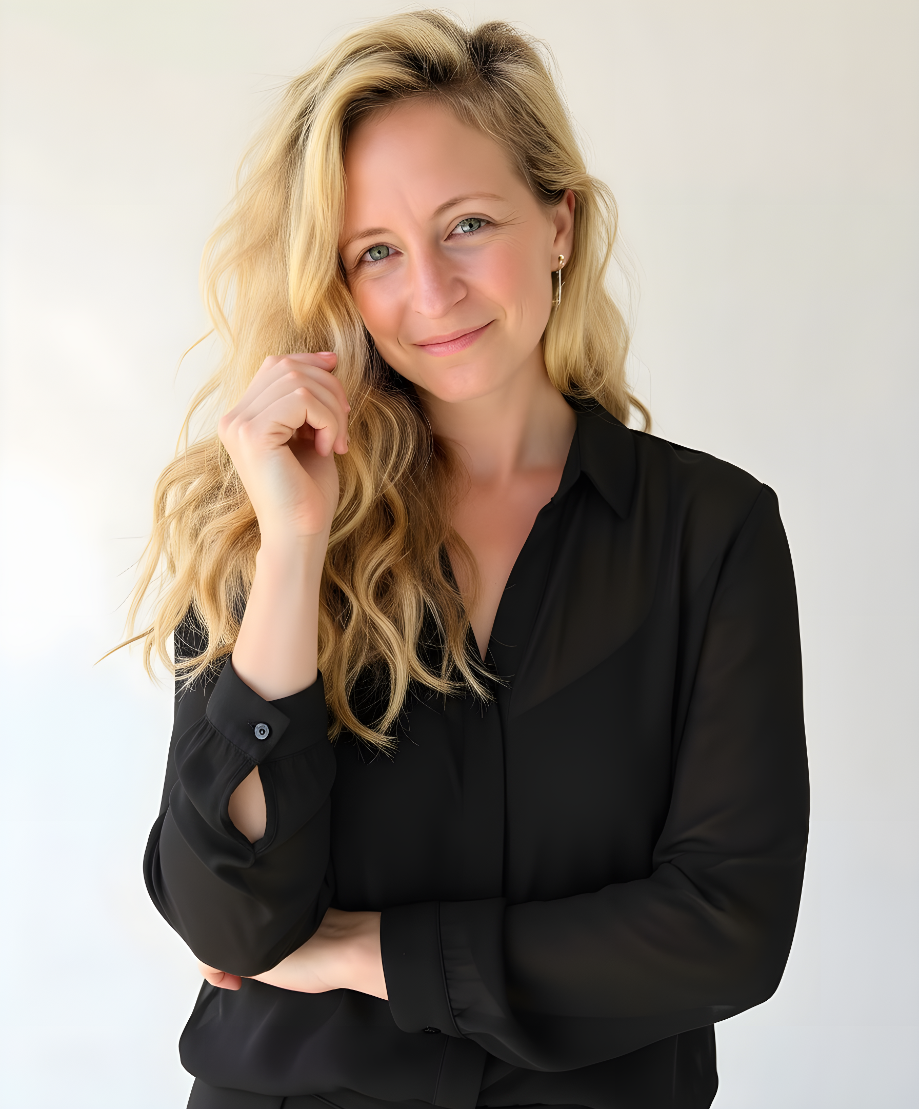 A woman with blonde hair wearing a black blouse, smiling softly with her hand touching her face, against a plain light background.