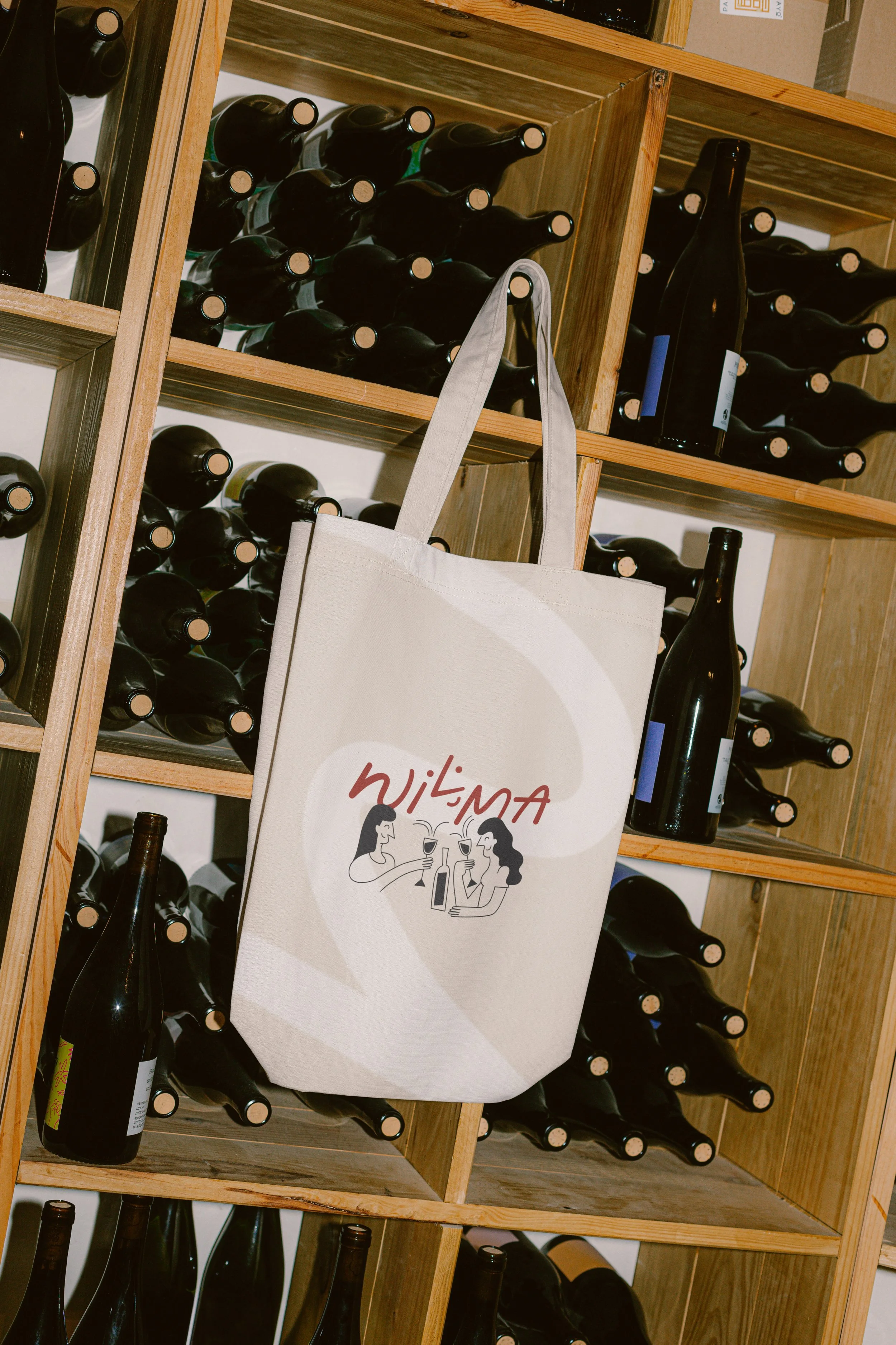 A beige tote bag with a graphic of two women holding wine glasses, hanging on a wine rack filled with bottles.