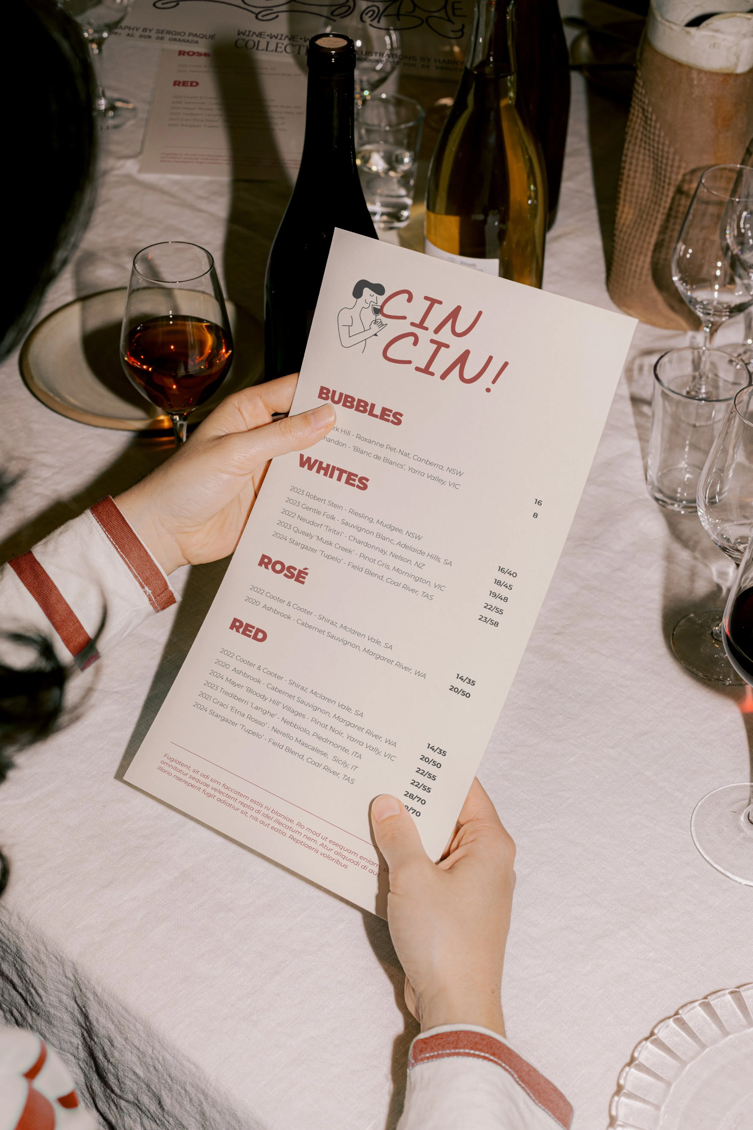 Person holding a wine menu titled 'CIN CIN!' at a dining table with various wine bottles, glasses, and a glass of red wine nearby.