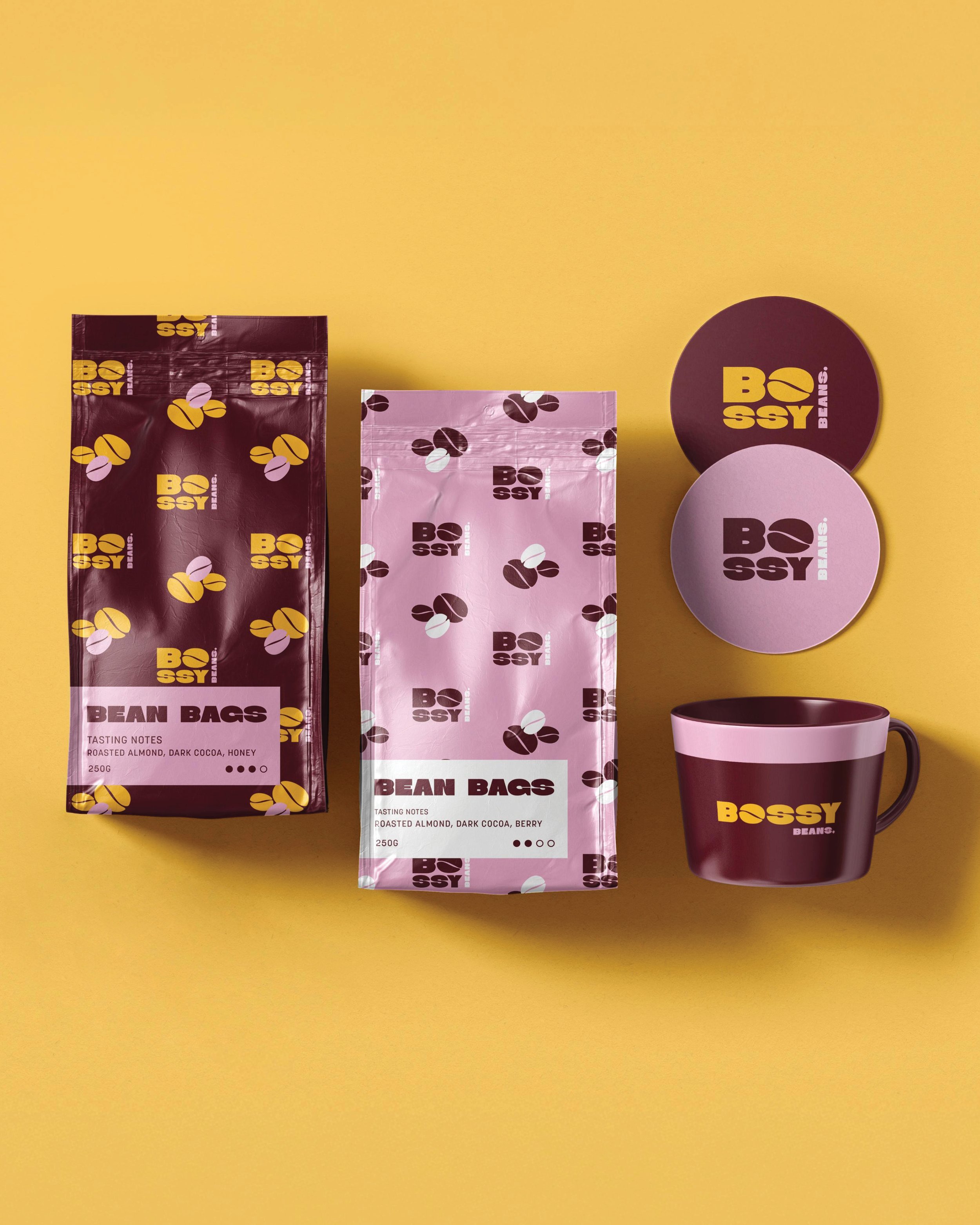 Packaging of Bossy Beans brand coffee, including two bags of bean coffee, two round coffee coasters and a coffee mug, all set on a yellow background.