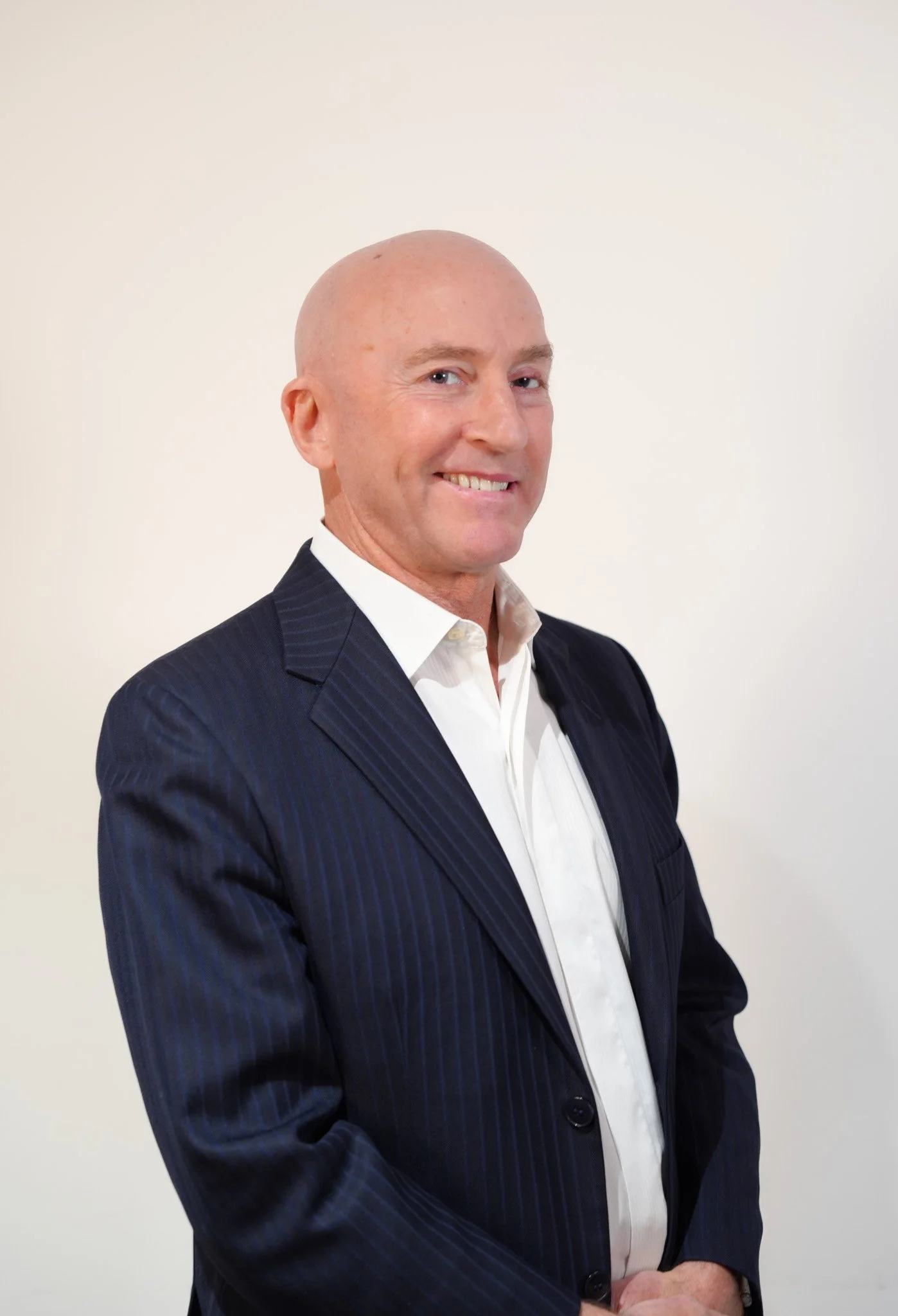 A man with a bald head wearing a dark pinstripe suit and white shirt, smiling and standing against a plain light background.