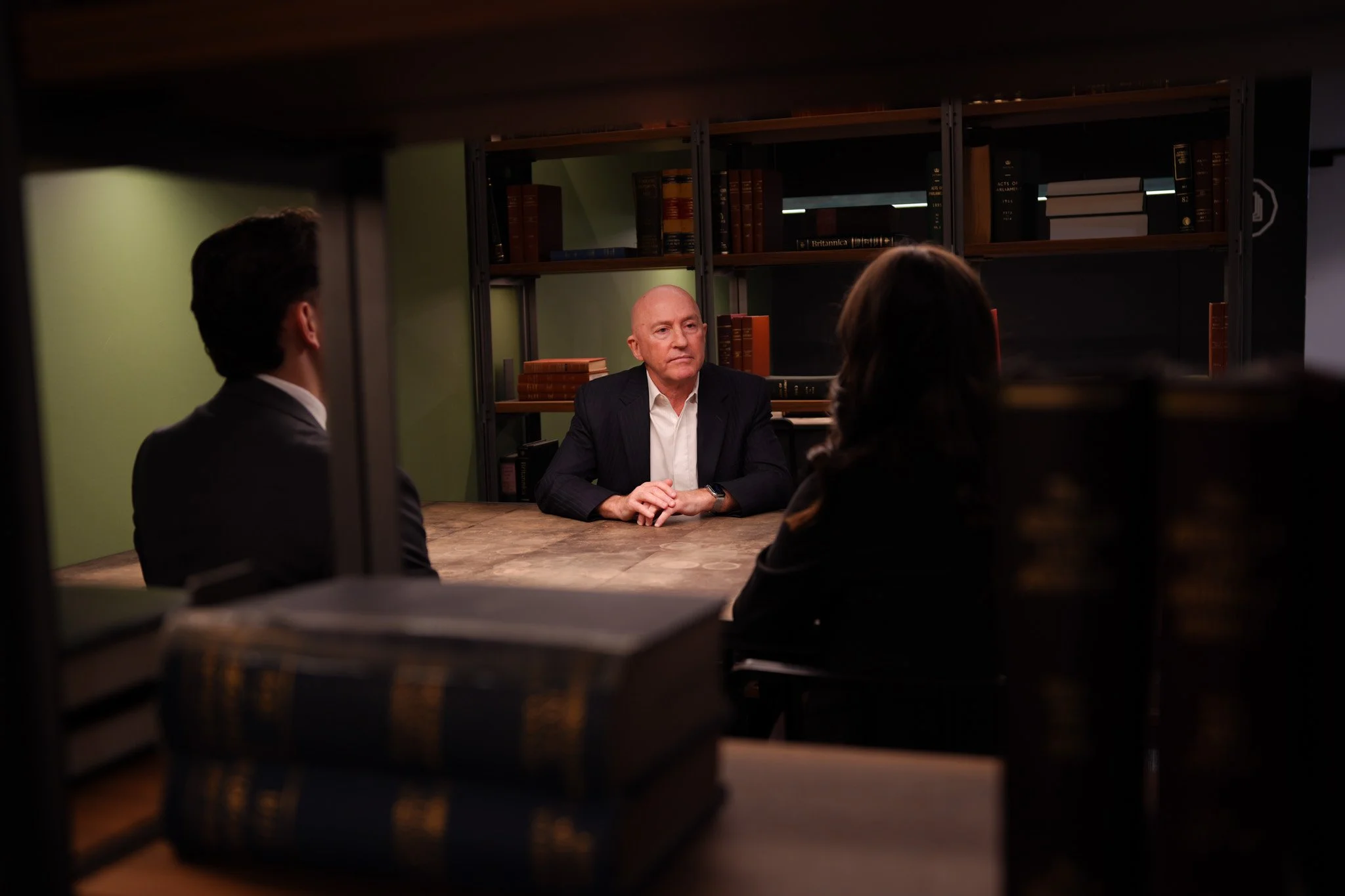 Three people having a discussion in a library or office, seated at a table behind bookshelves, with a man in a dark suit facing two people on the other side.