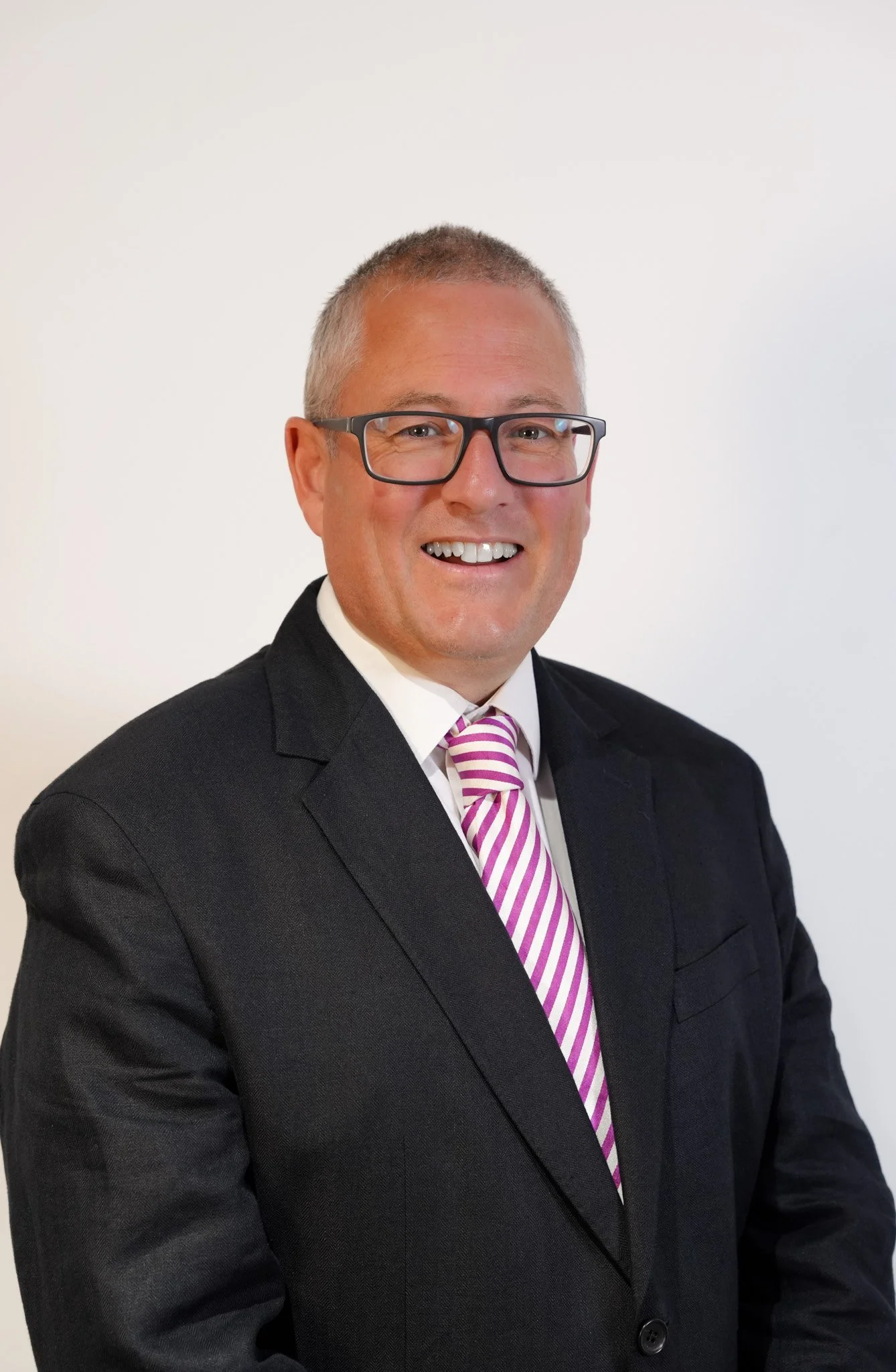 A man in a black suit, white shirt, and pink and white striped tie smiling at the camera against a plain light background.
