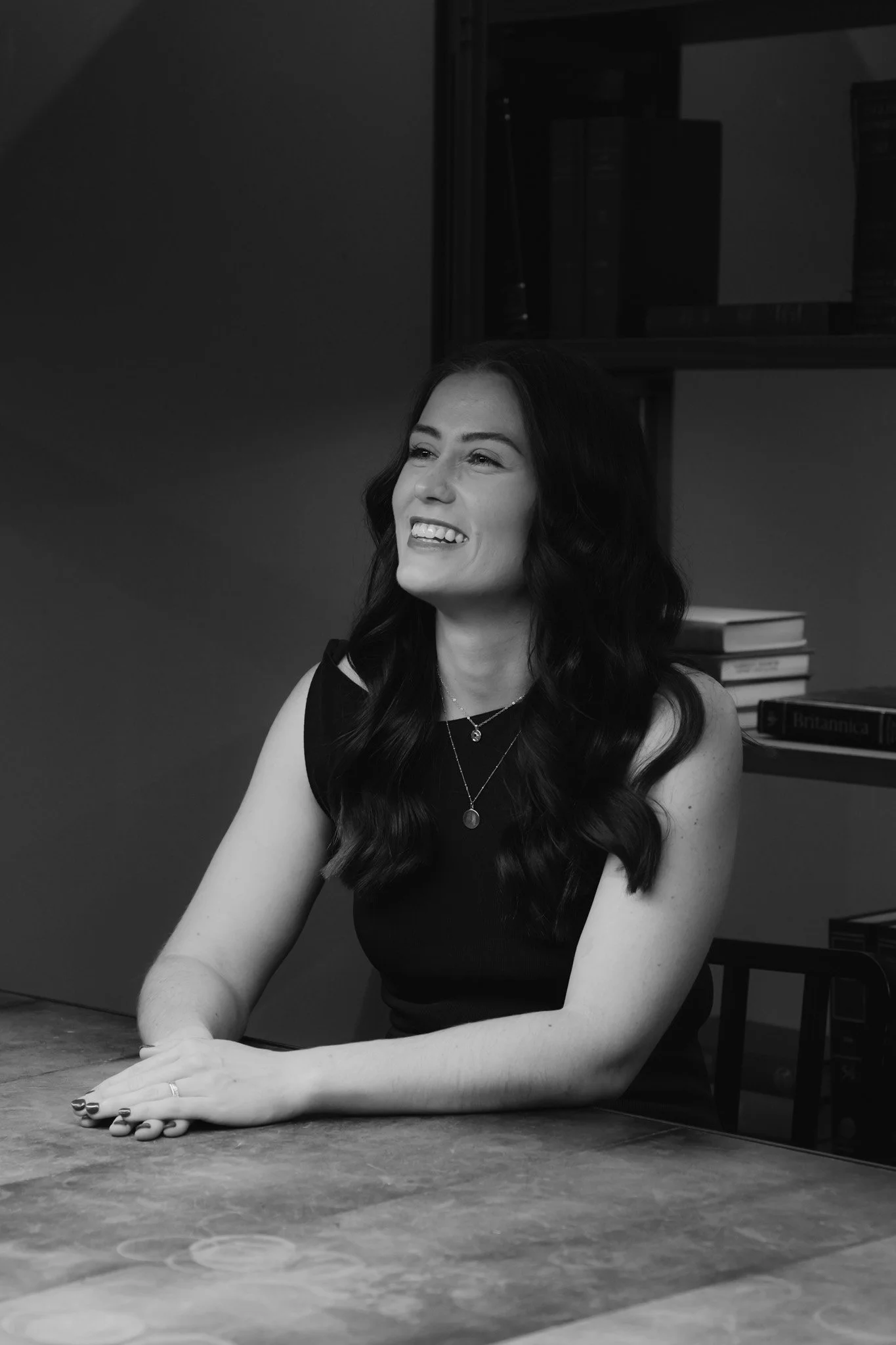 A woman with dark wavy hair smiling and sitting at a table in an indoor environment, with books and a dark bookshelf in the background.