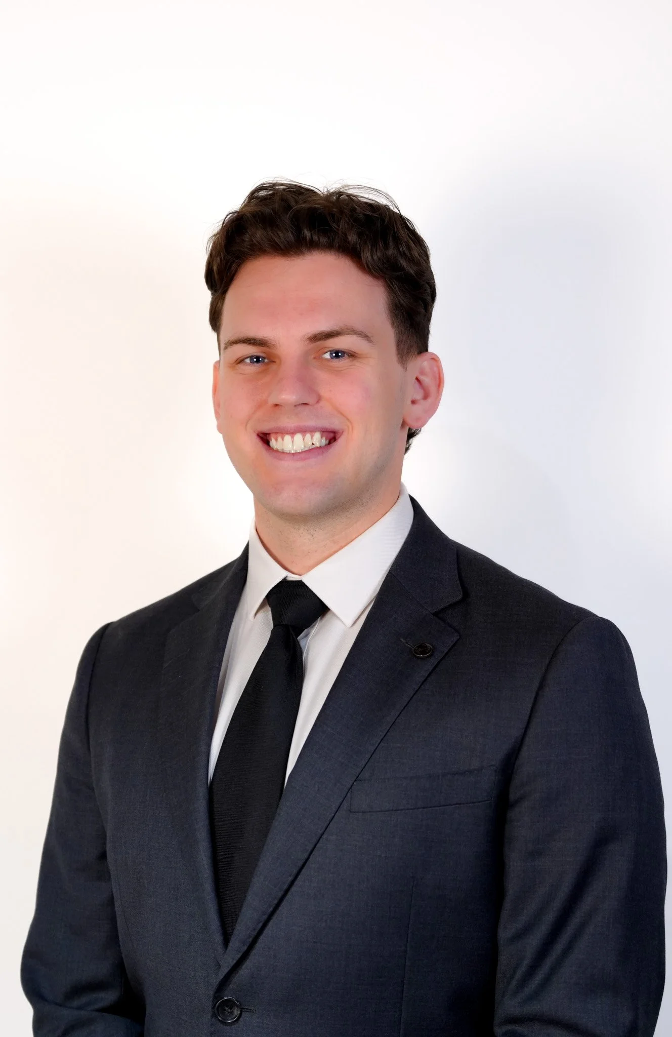 A young man with dark hair wearing a black suit, white shirt, and black tie posing against a white background.