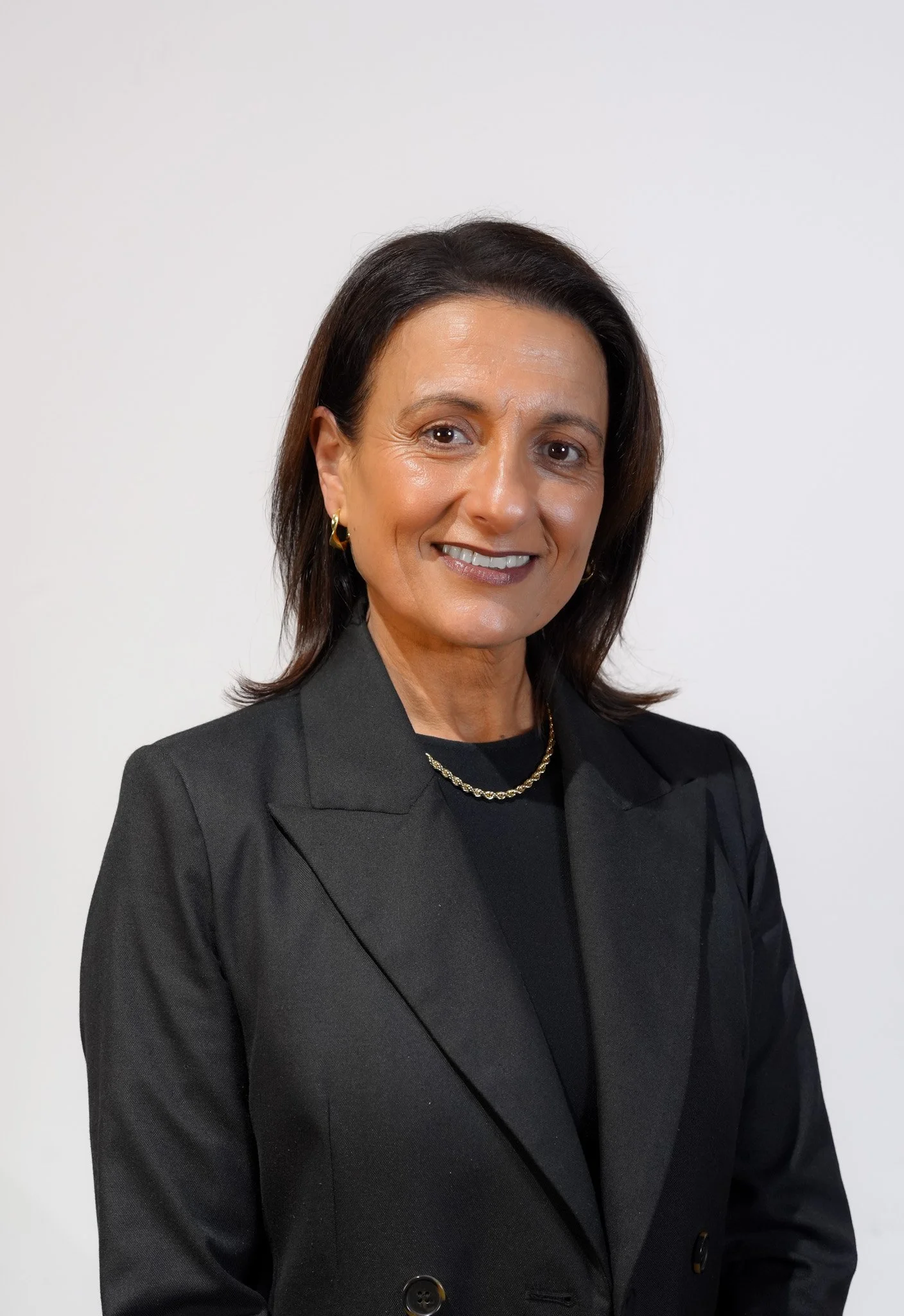 A woman with shoulder-length brown hair is smiling and wearing a black blazer, gold hoop earrings, and a gold necklace, standing against a plain white background.