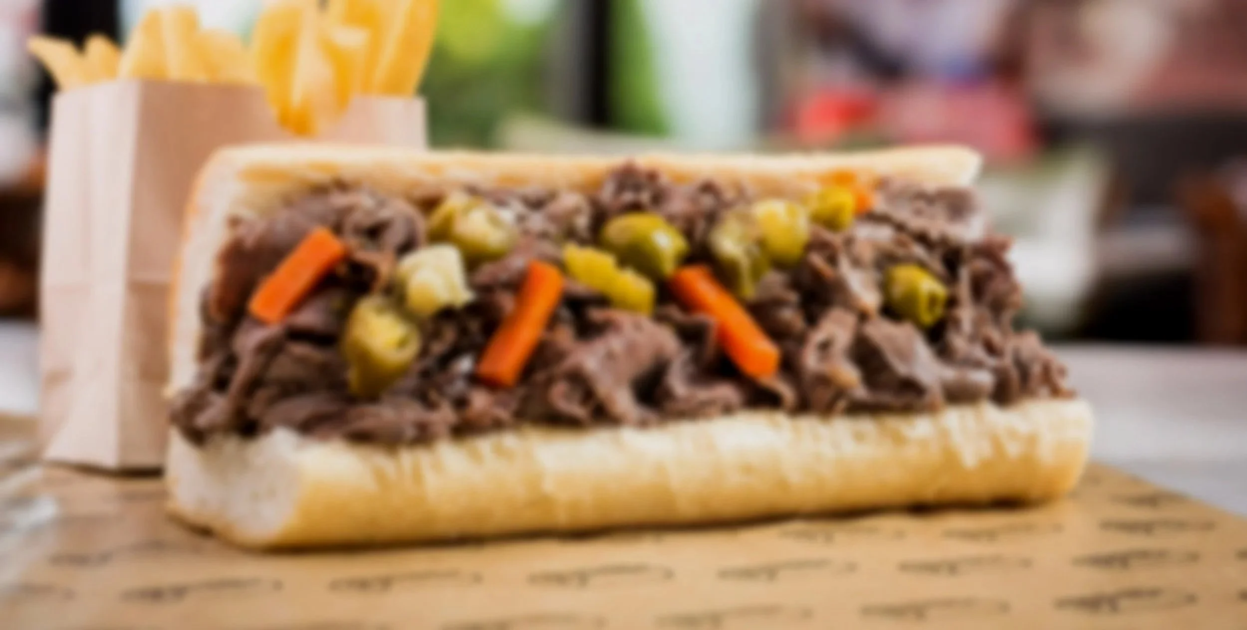 Philly cheesesteak sandwich with shredded beef, chopped pickles, and sliced carrots on a hoagie bun, served with French fries in a paper container.