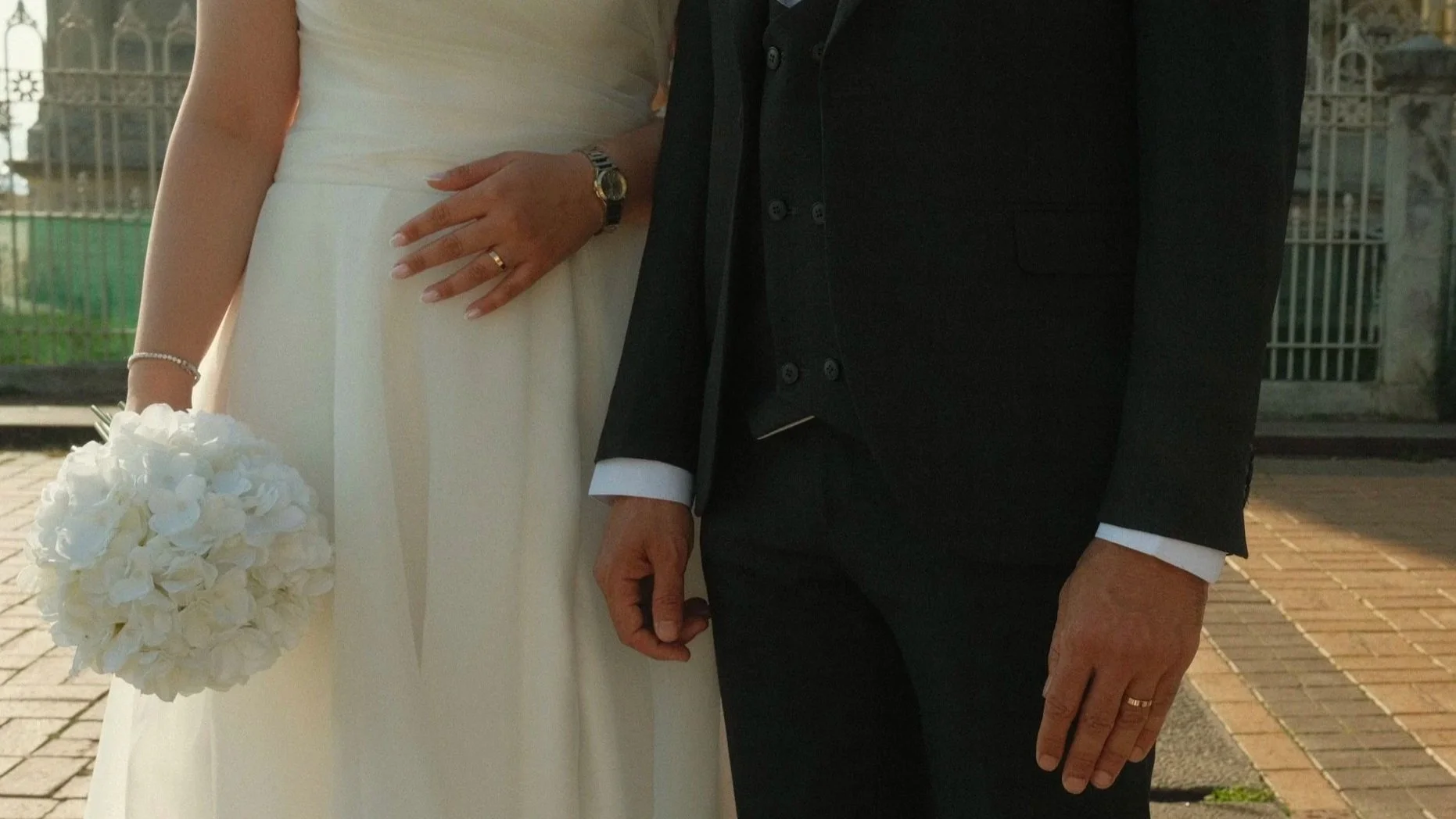 Close-up of a bride and groom standing side by side outdoors. The bride is wearing a white dress, a bracelet, a watch, a ring, and holding a bouquet of white flowers. The groom is dressed in a black suit with a white shirt, and a ring on his finger.