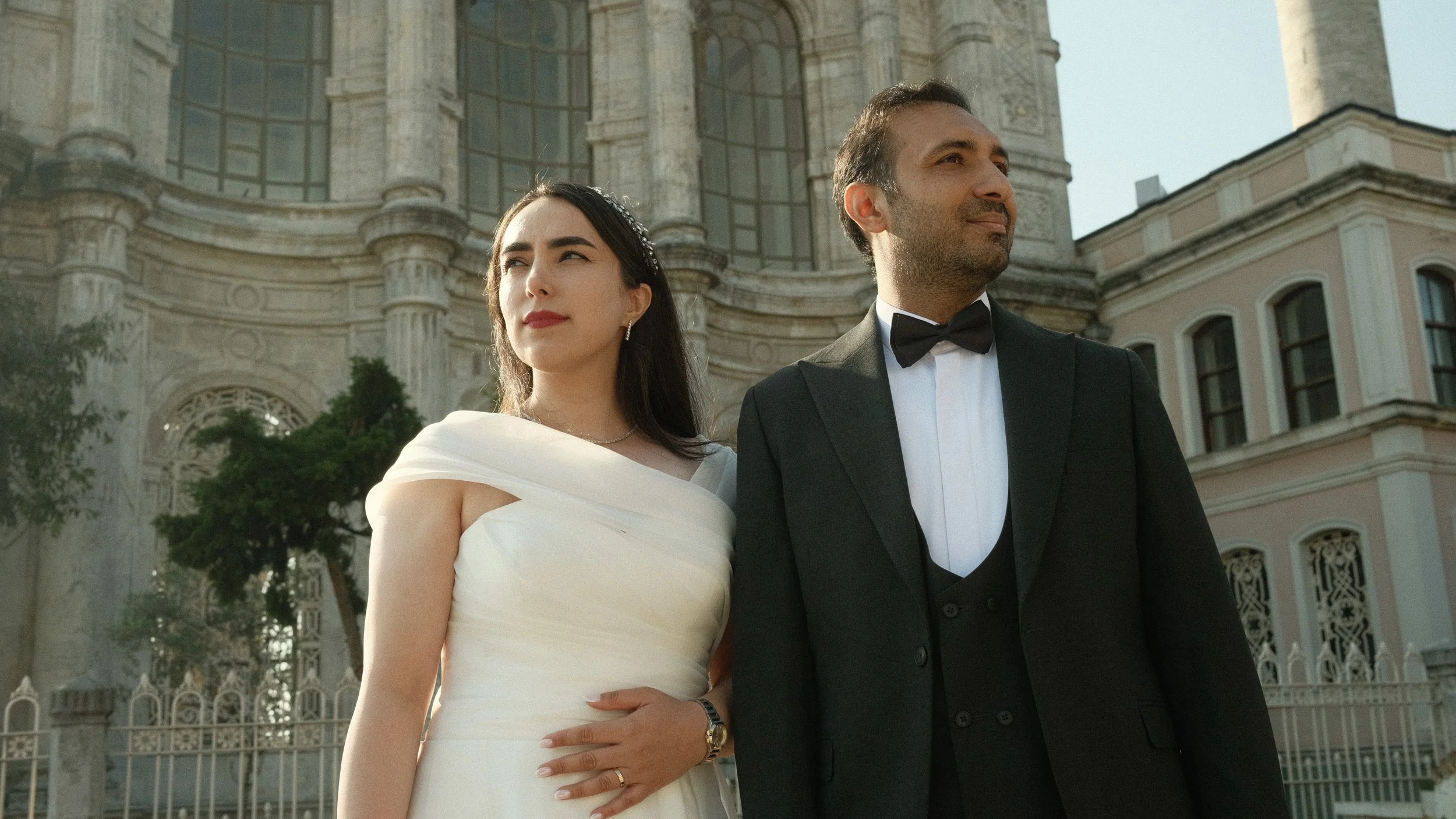 A bride and groom in formal attire standing outdoors in front of a historic building with large arched windows.
