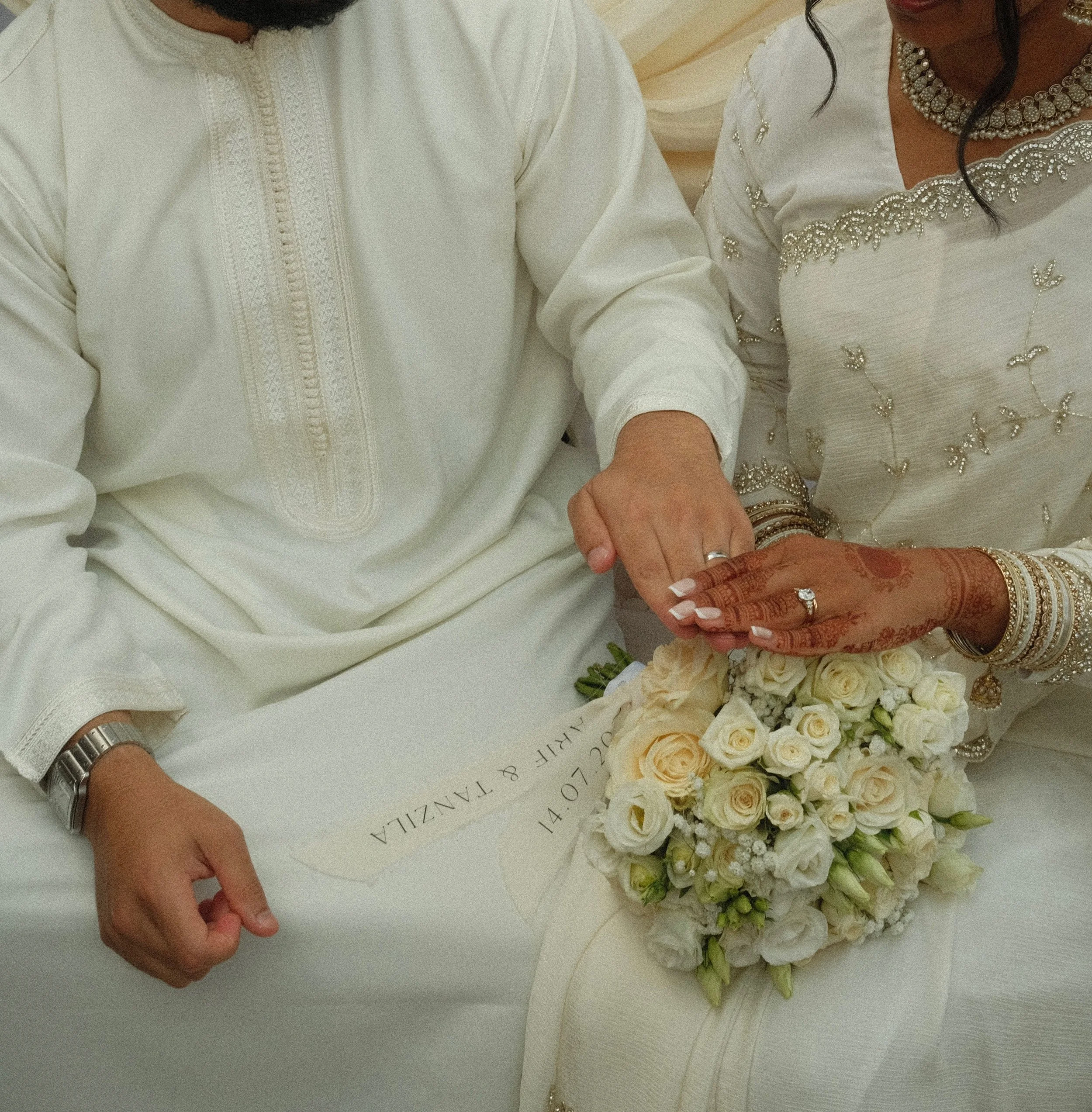Close-up of two people holding hands, one wearing traditional white clothing with detailed embroidery, and the other wearing ornate jewelry and a cream-colored dress with silver embellishments, surrounded by a bouquet of white roses.