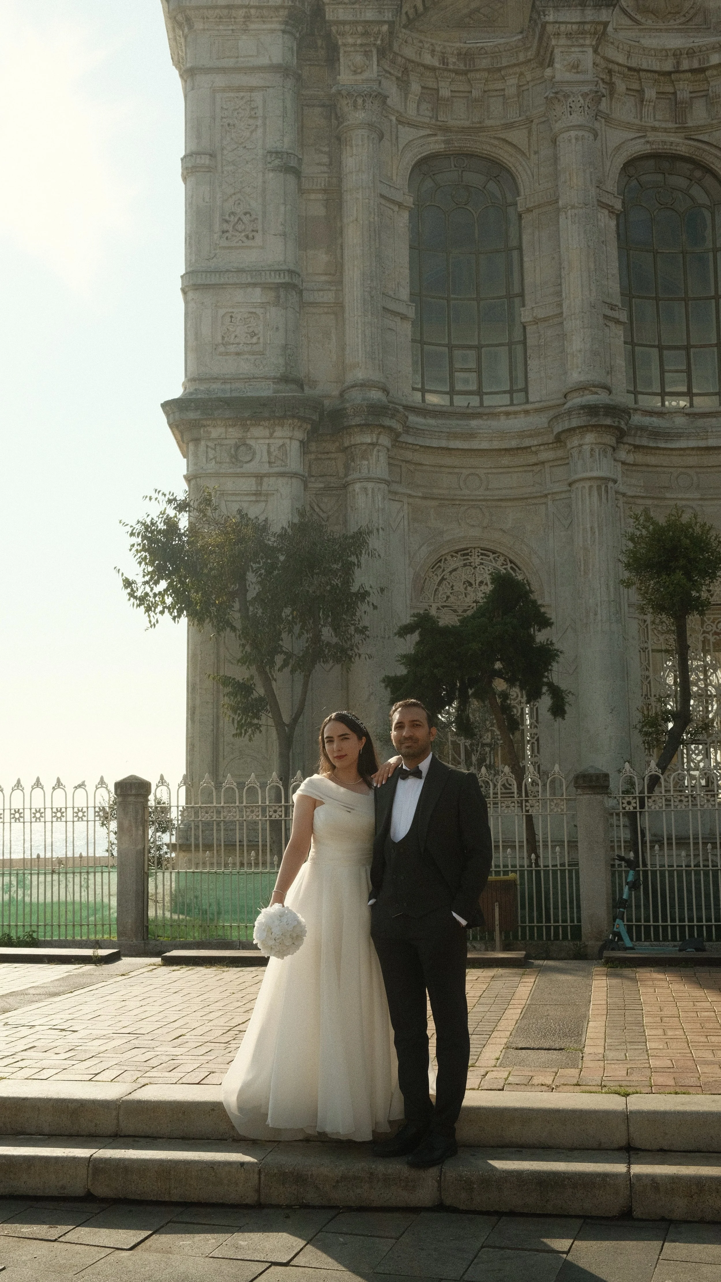 A bride in a white wedding dress holding a bouquet and a groom in a black tuxedo posing in front of a historic building with large windows and ornate architectural details.