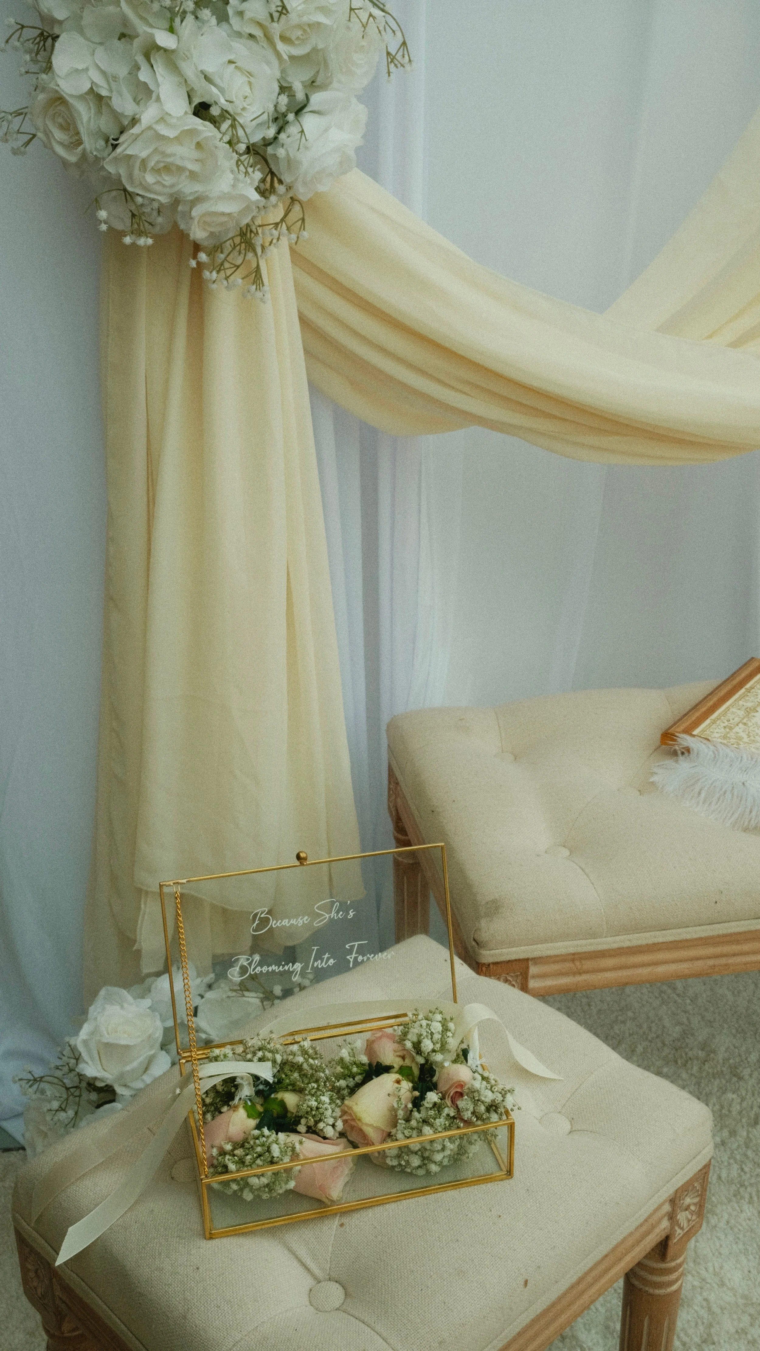 A wedding or event decoration with a floral arrangement, cream-colored drapes, a vintage-style chair, and a glass box with flowers and a sign that reads "Because She's Blooming Into Forever".