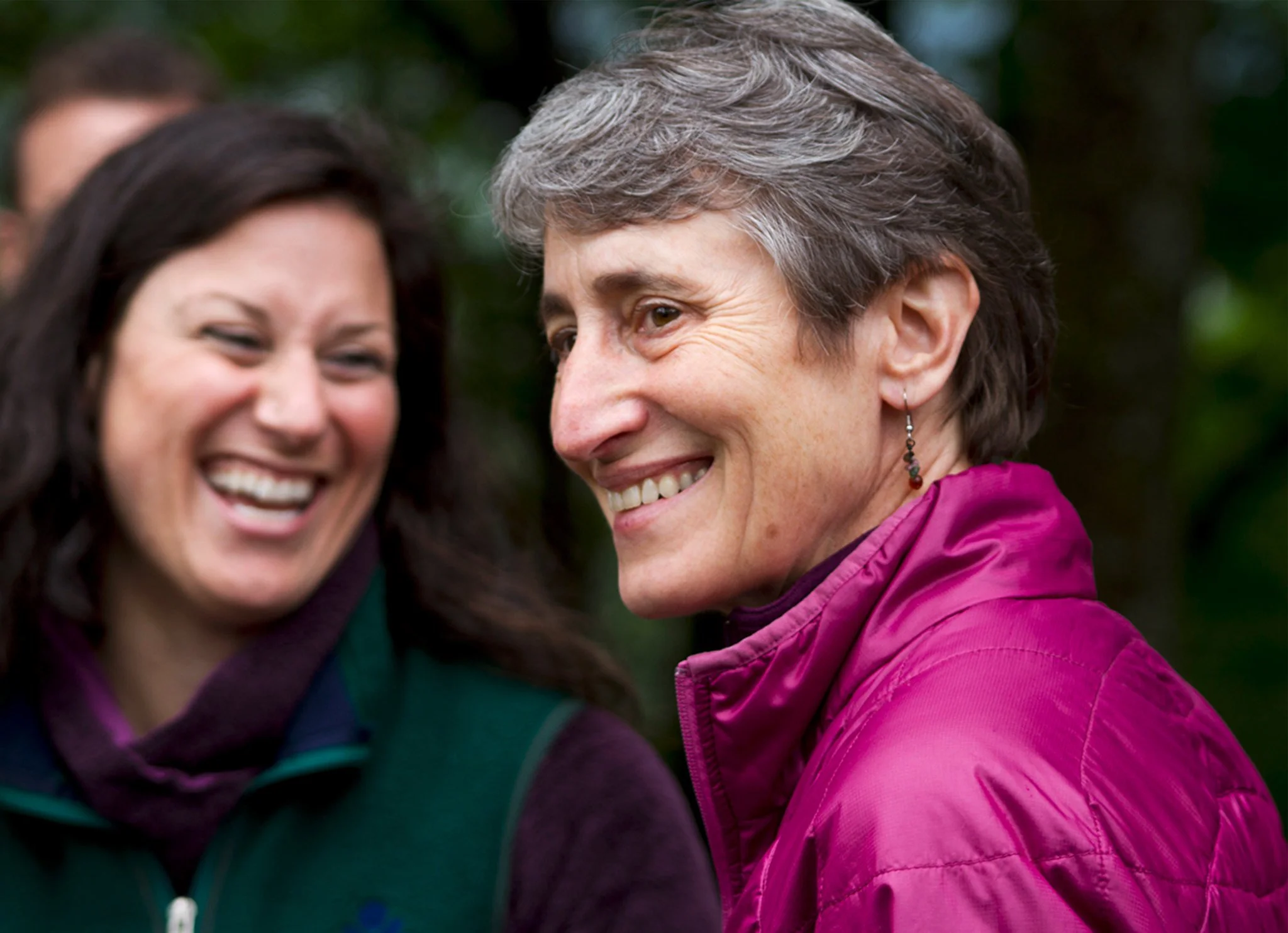 Two women smiling and laughing outdoors, with a blurred natural background.