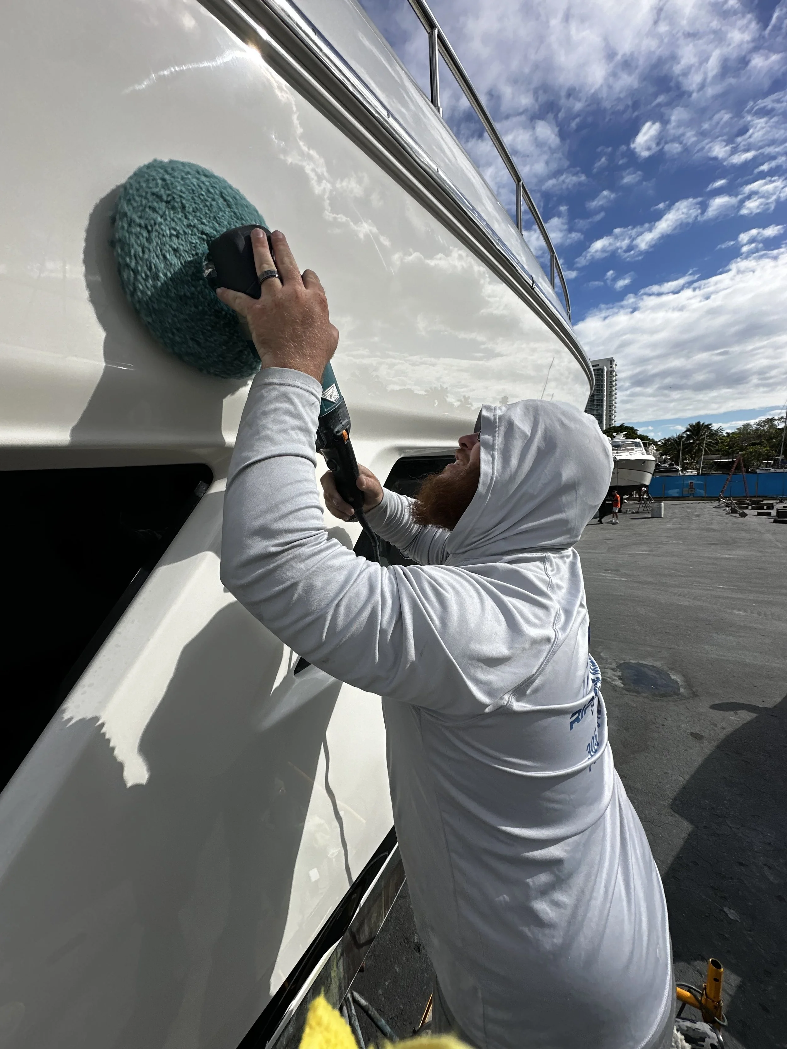 A person wearing a white hoodie polishing a boat's hull with a green scrubbing pad using a power tool at a marina.