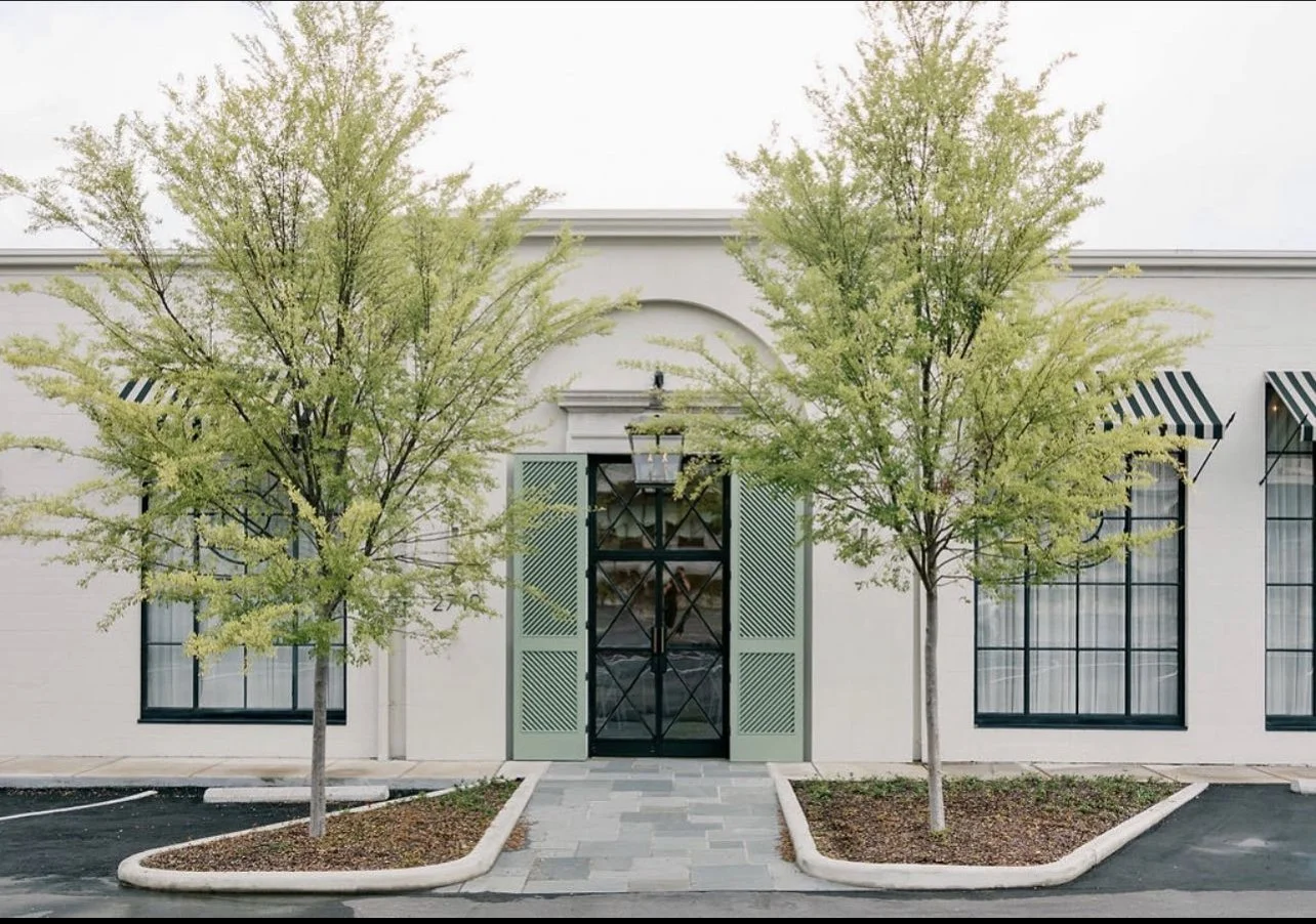 White building with black and white striped awnings, two trees in landscaped beds, and a central glass door with green shutters.