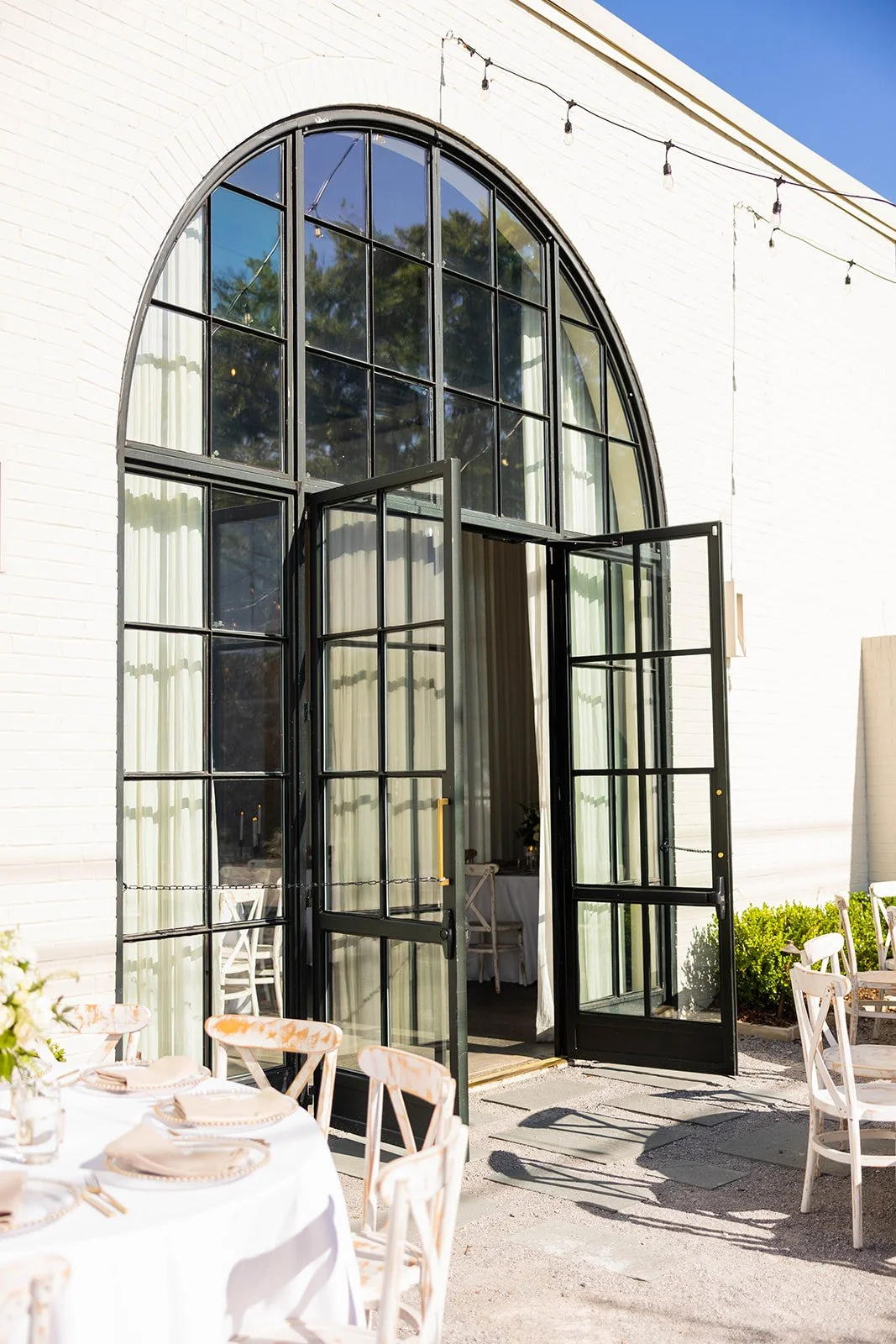 Open black-framed glass door leading to a decorated outdoor dining area with white tablecloths and chairs, set for a meal, adjacent to a white brick building with large arched windows and string lights overhead.