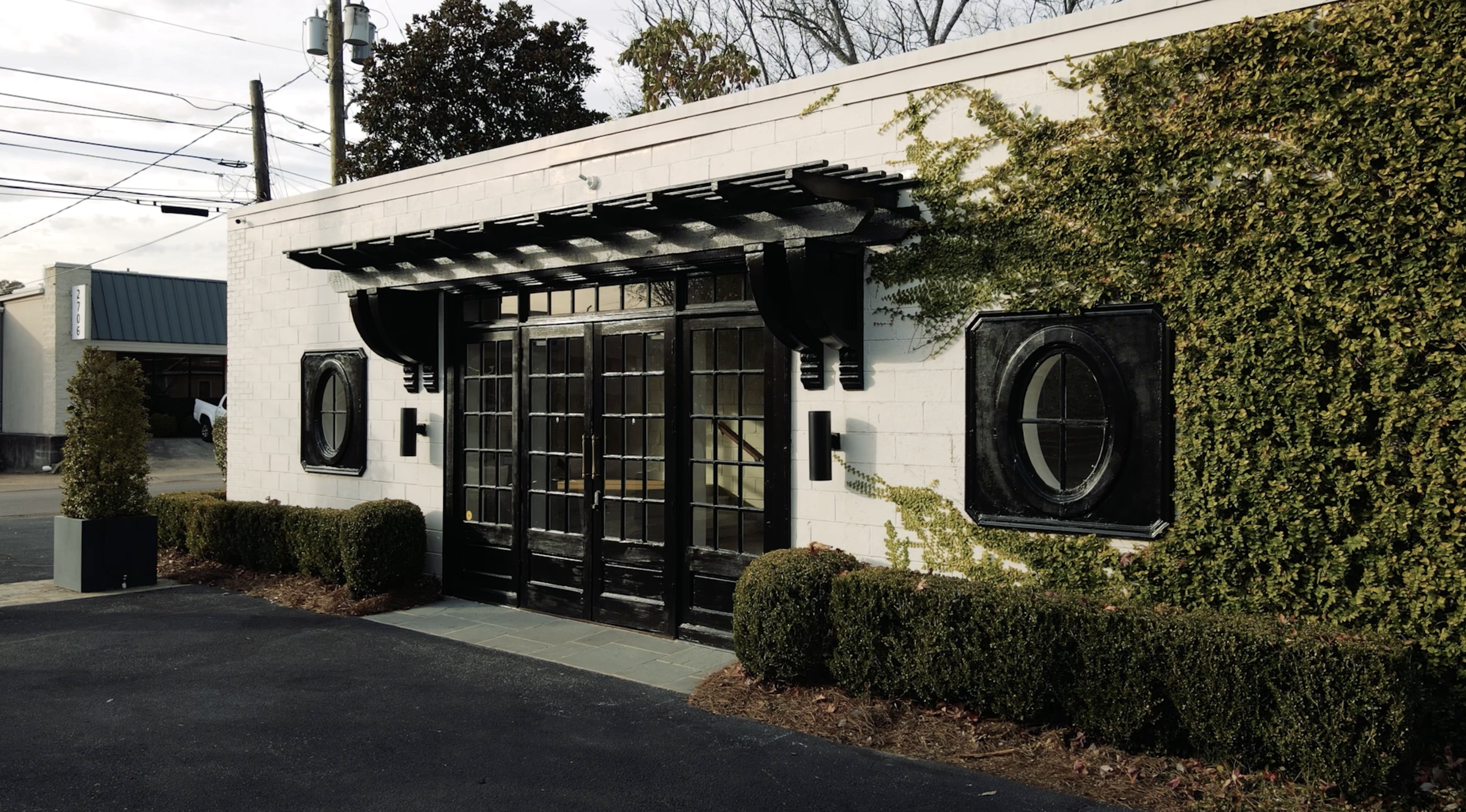 Exterior of a modern building with a white brick wall, black framed glass double doors, black decorative elements, two circular black window frames, green ivy on the right, and trimmed bushes along the front.