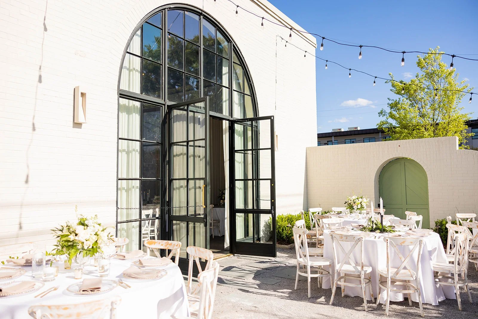 Outdoor wedding reception setup with round tables covered in white tablecloths, decorated with floral centerpieces, set up outside a building with large arched window and black open doors, string lights overhead, and a brick wall with a green door in the background.