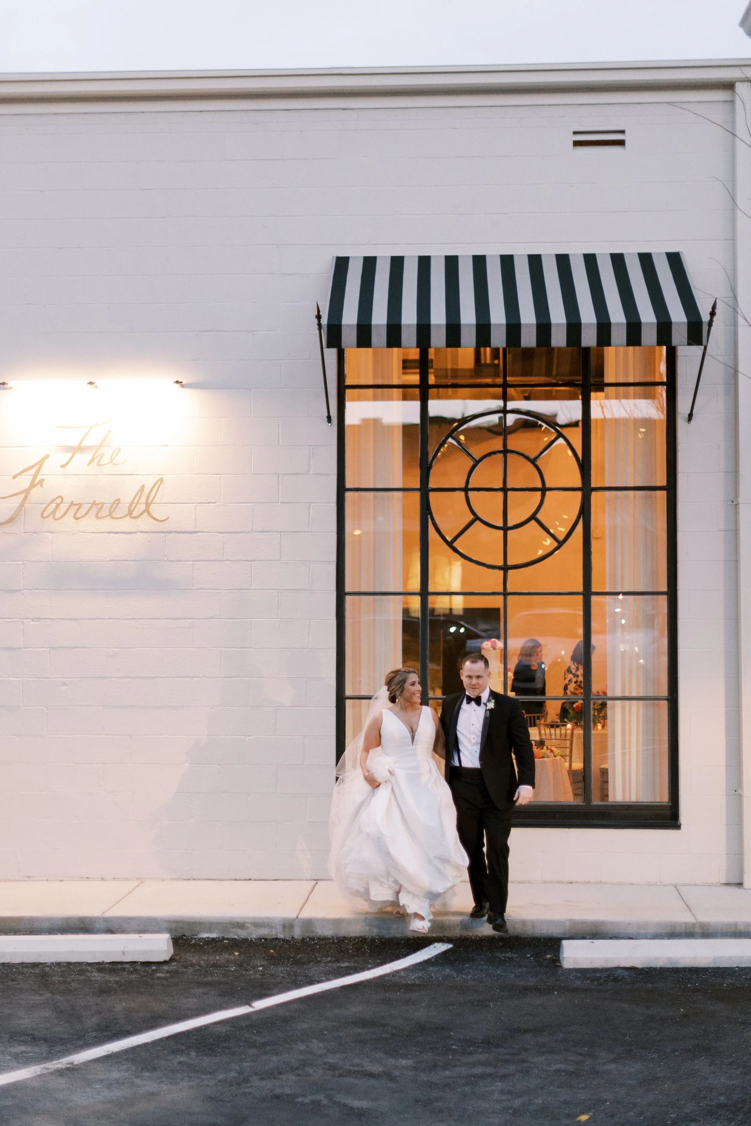 A newly married couple walking out of a building named 'The Farrell,' with the bride in a white wedding dress and the groom in a black tuxedo, through a large window with orange curtains inside the building.