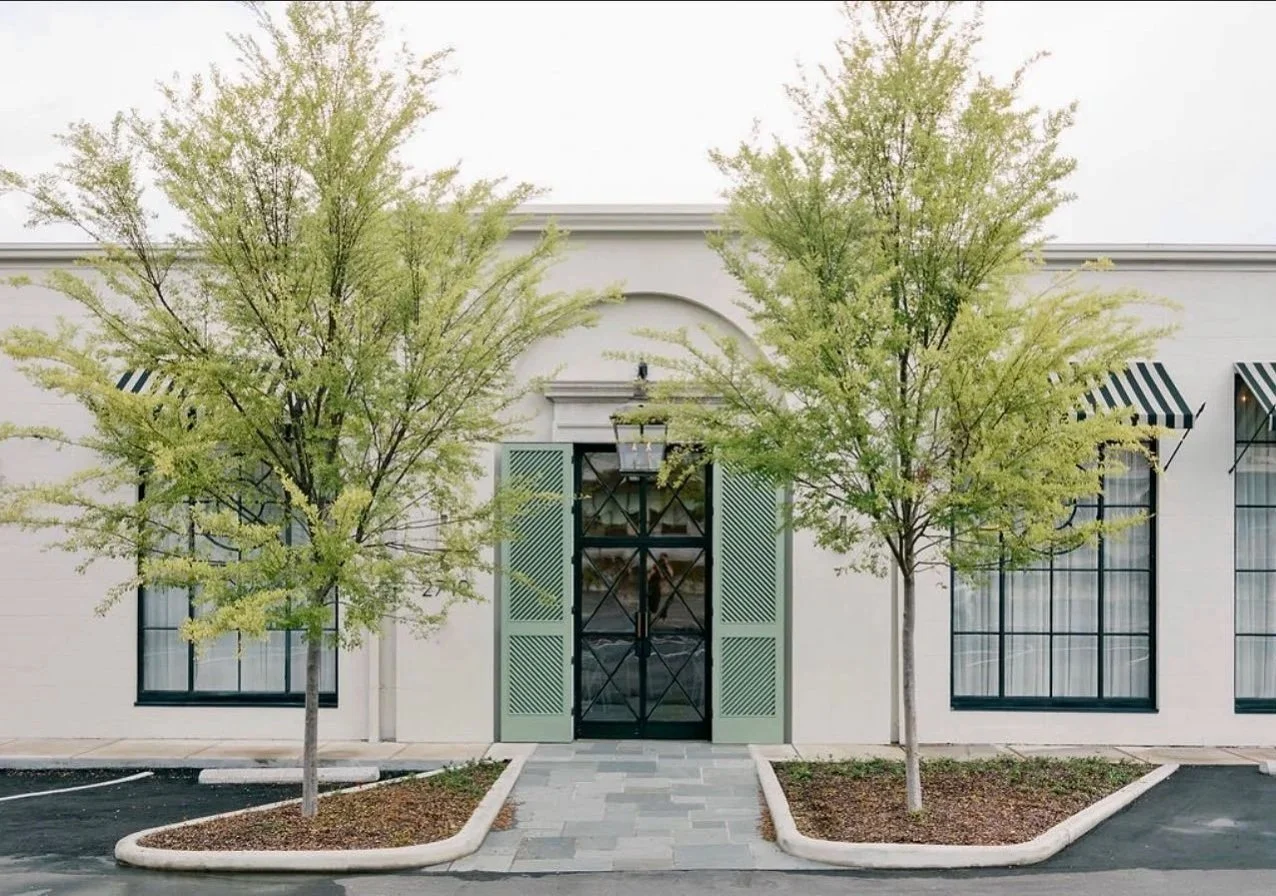 White building with green shutters and striped awnings, two trees in front, and a paved walkway leading to the entrance.
