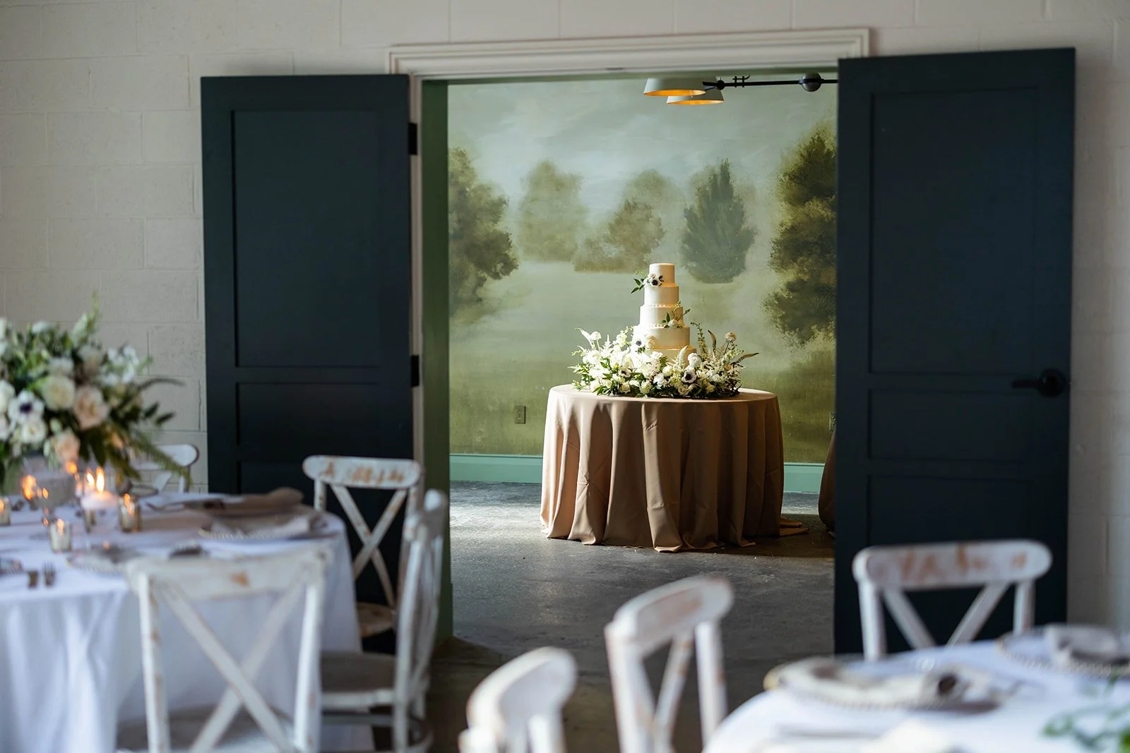 A wedding cake on a round table decorated with white flowers, seen through open black doors in a decorated event hall with tables and chairs.