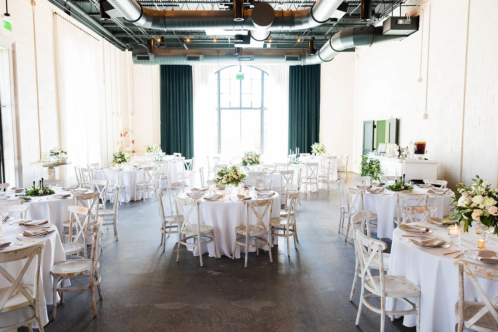 Elegant event space decorated with round tables covered in white tablecloths, set with beige napkins, tableware, and floral centerpieces, featuring a large arched window with green curtains and industrial ceiling with exposed ducts.