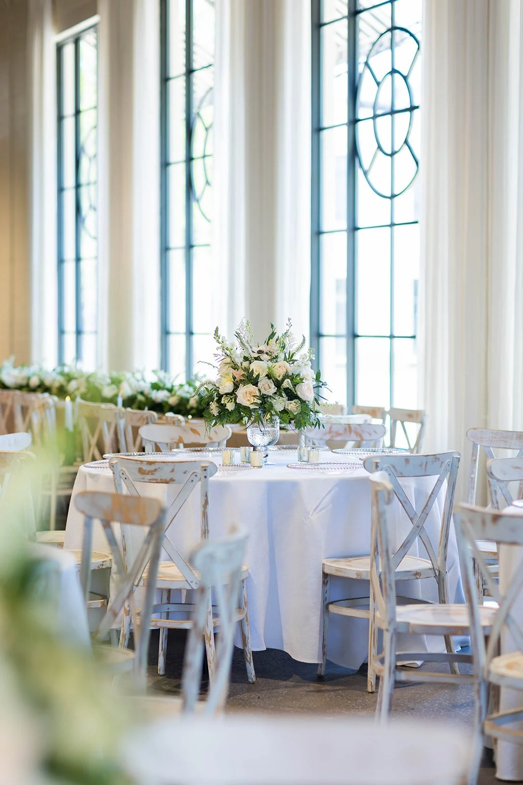 Elegant event setup with a round table draped in a white tablecloth, floral centerpiece featuring white roses and greenery, surrounded by white wooden chairs, large windows with arched panes, and sheer curtains in a bright, airy room.