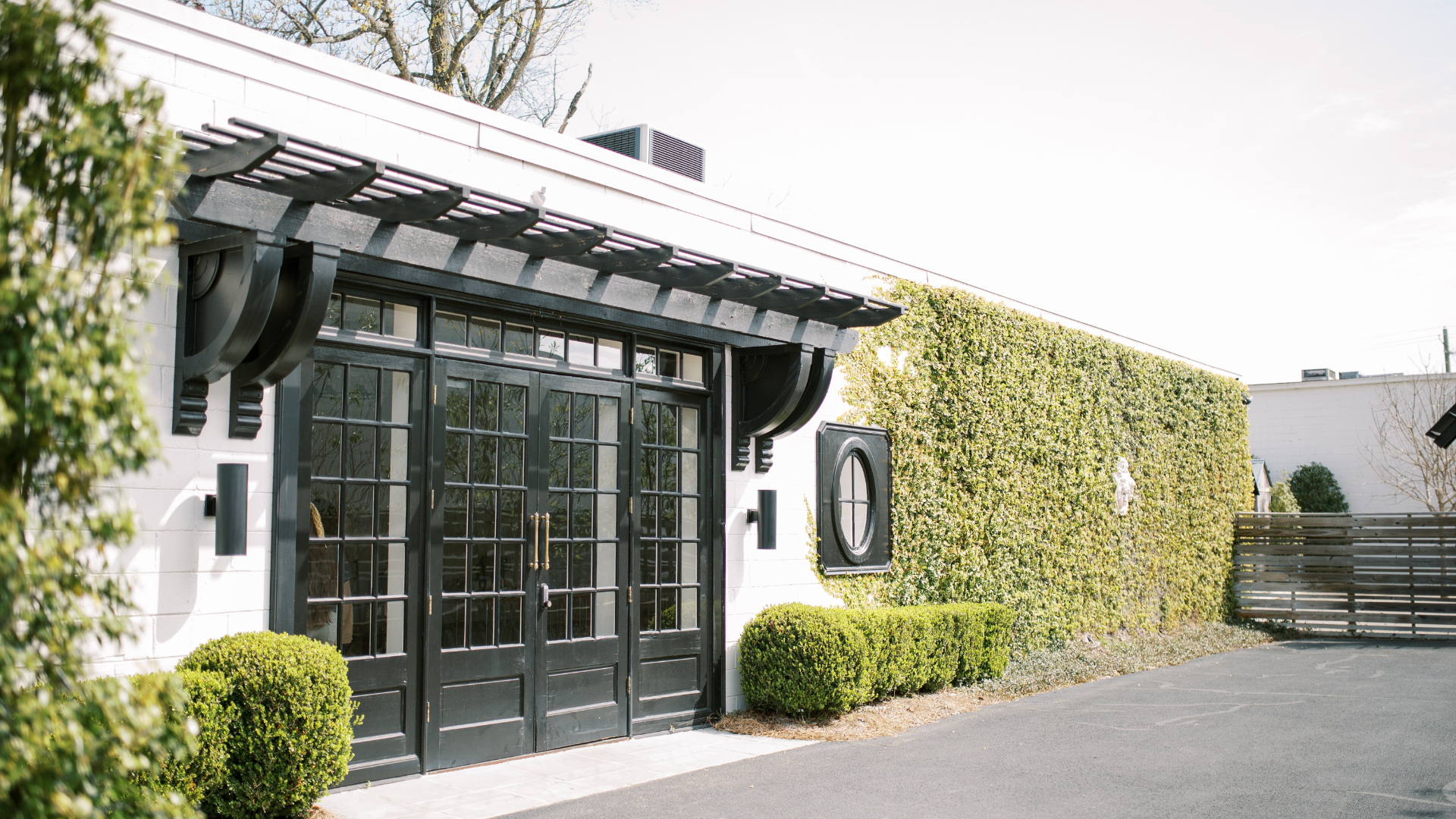 Exterior of a building with black glass double doors, a white wall, and a tall hedge. There are black light fixtures and a decorative round window on the wall. A small shrub is in front of the wall and a paved driveway nearby.